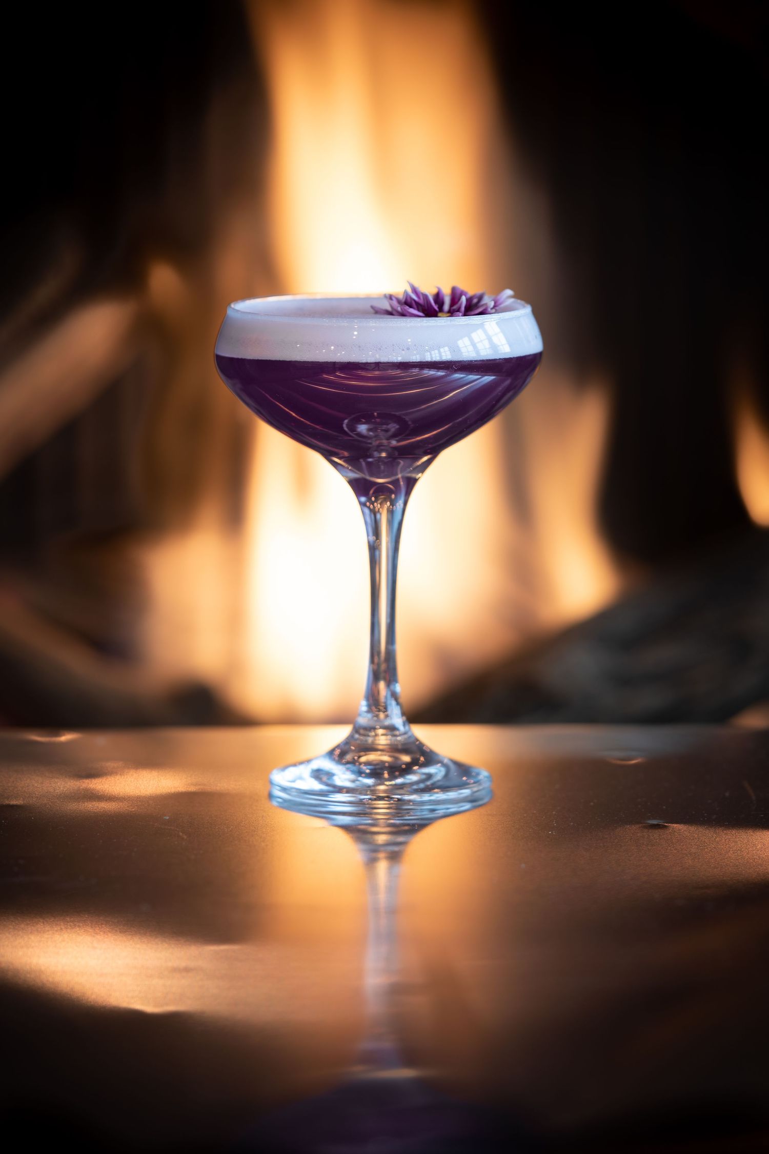 Elegant purple cocktail in coupe glass garnished with flower petals against warm glowing fireplace background.