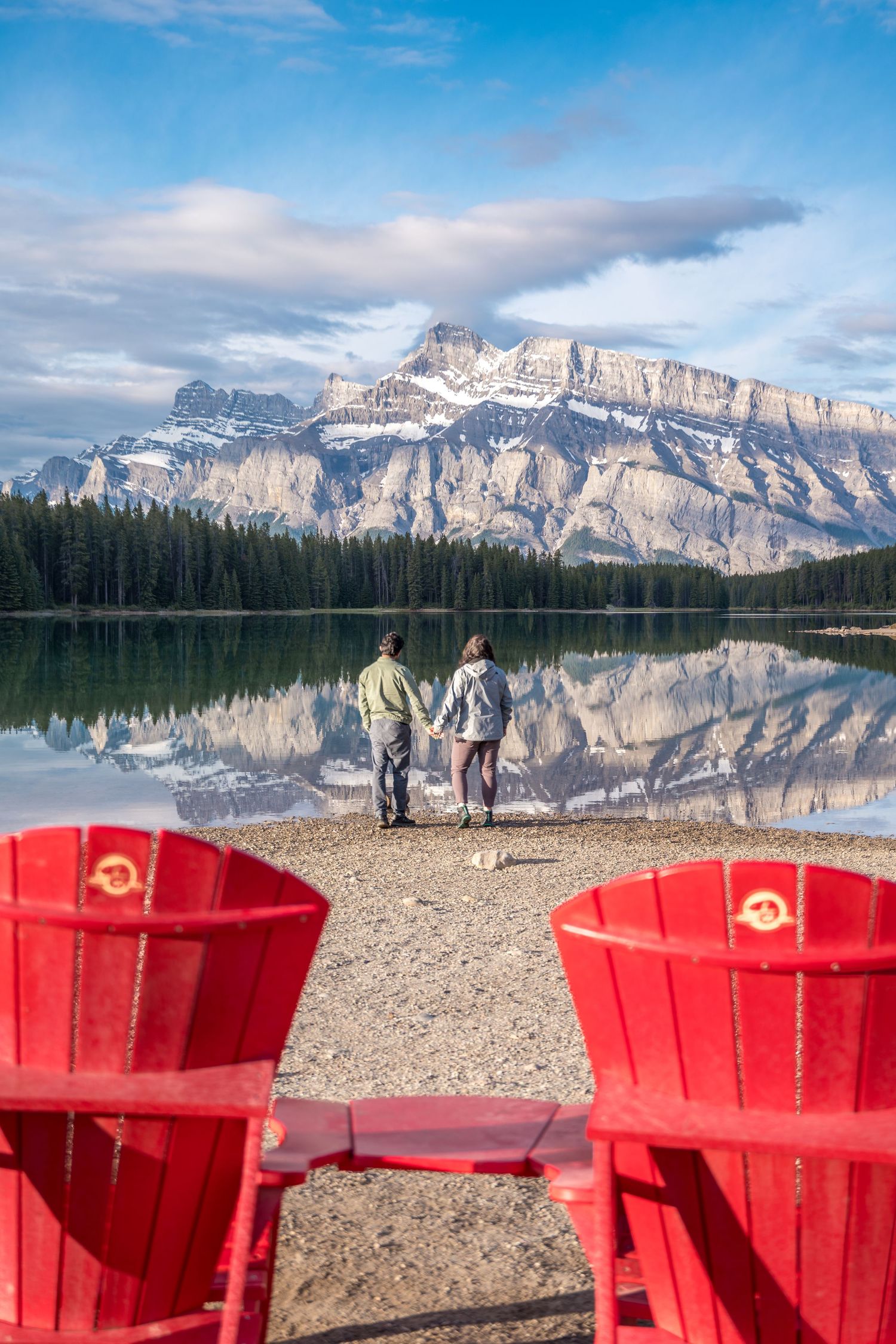 Two red Adirondack chairs overlook a couple at Two Jack Lake with Mount Rundle reflected in calm waters, Banff, Canada.