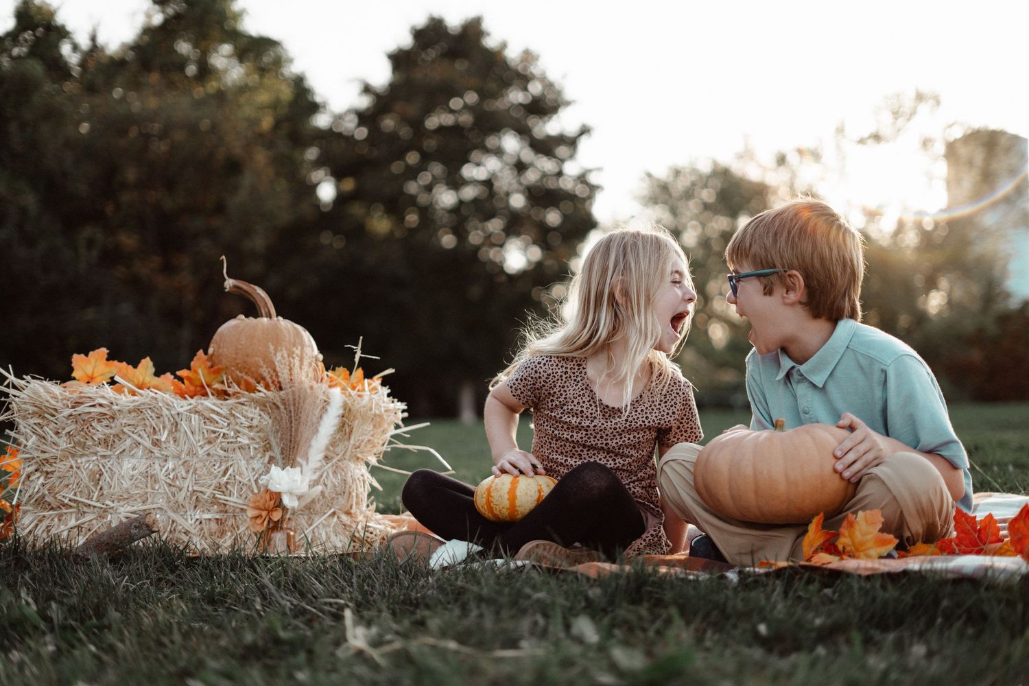 Two children sit on a blanket surrounded by pumpkins and hay bales during a fall outdoor photo session.