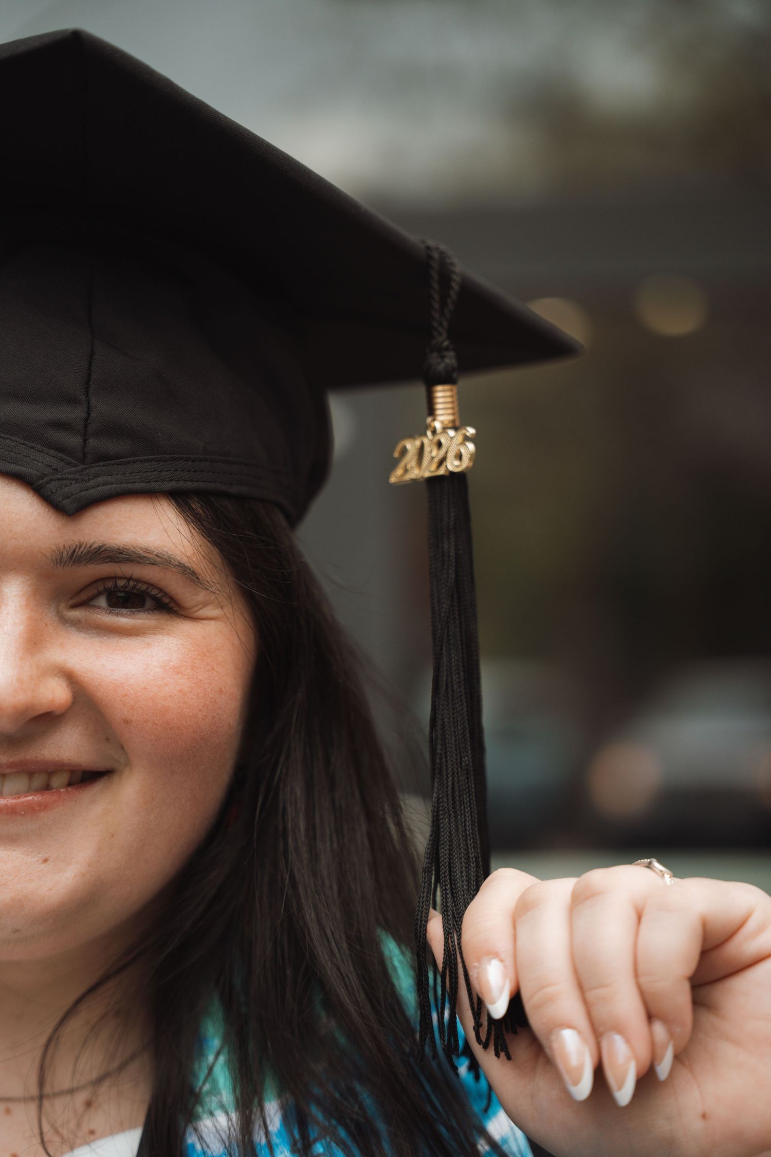 A smiling graduate holds her black mortarboard cap with a gold 2025 tassel close to the camera.