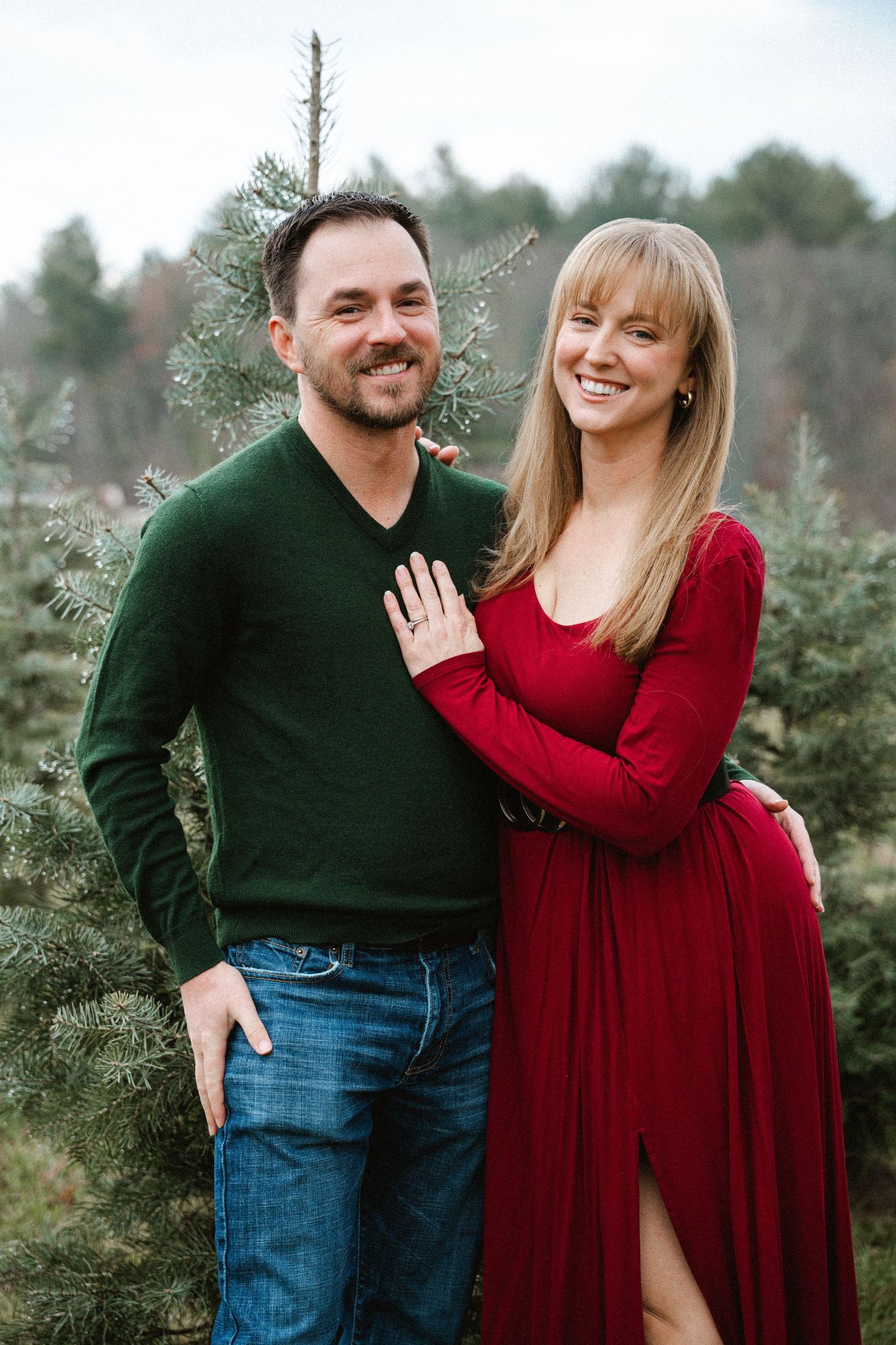 A smiling couple poses together among Christmas trees, wearing festive red and green holiday outfits.