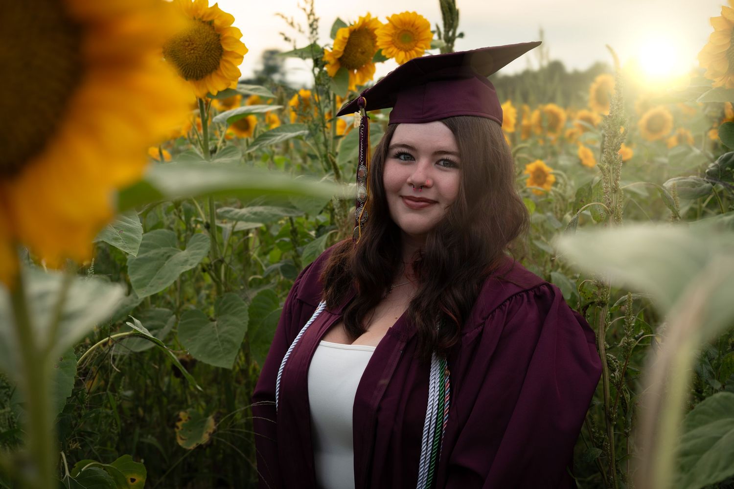 Smiling graduate in maroon cap and gown poses among bright sunflowers at sunset.