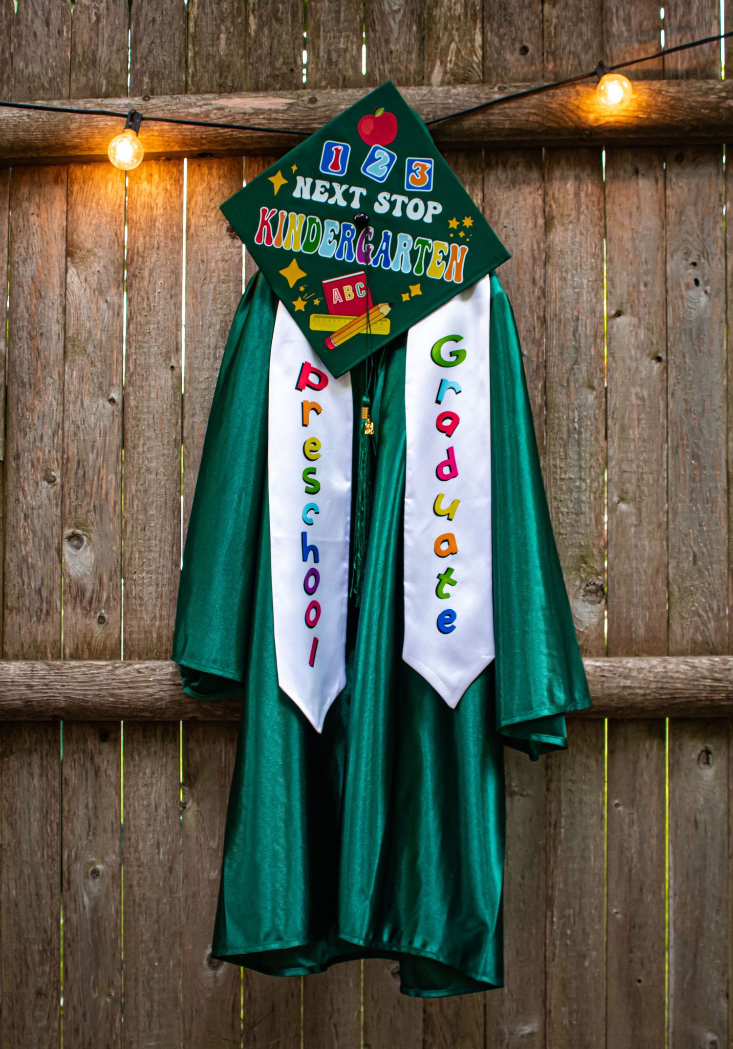 Green preschool graduation gown with cap reading Next Stop Kindergarten on wooden fence.
