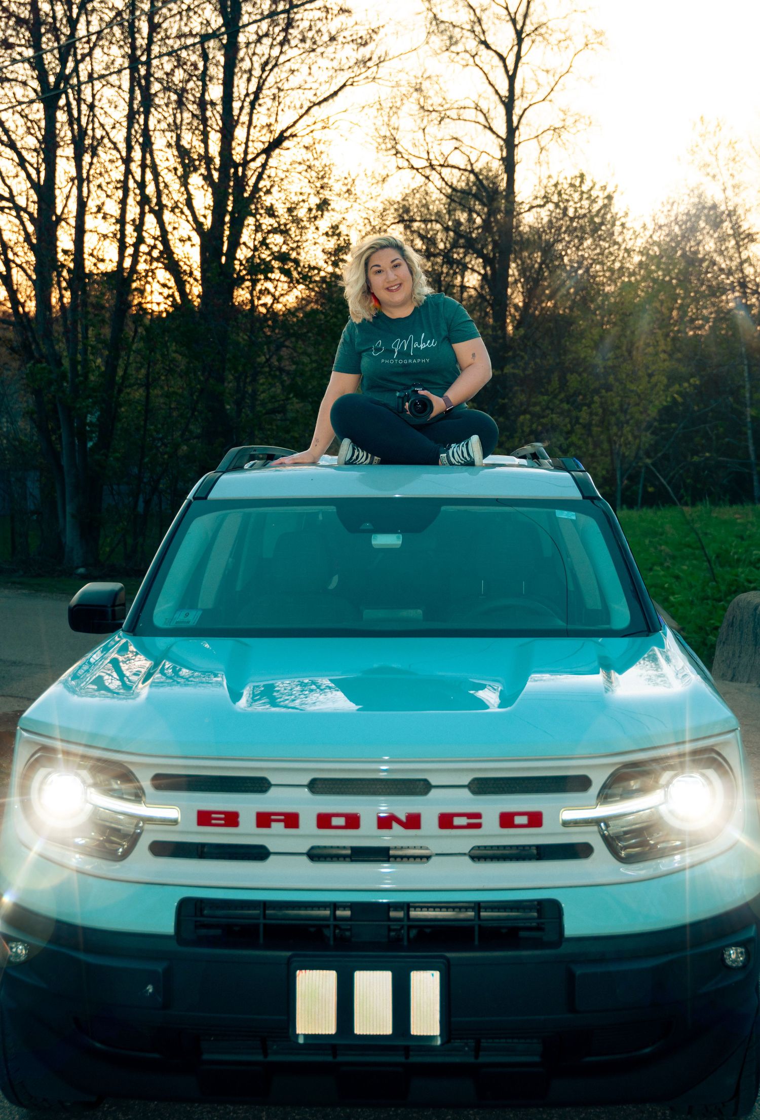 Woman sitting on hood of teal Ford Bronco SUV at sunset with headlights on.