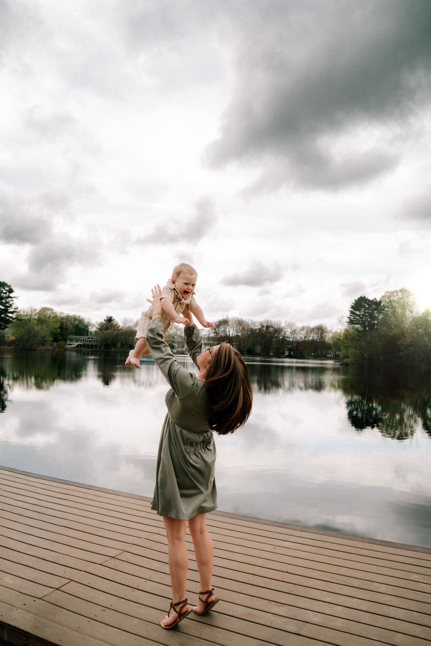 Joyful mother lifting laughing baby high in the air on a dock under dramatic cloudy skies.