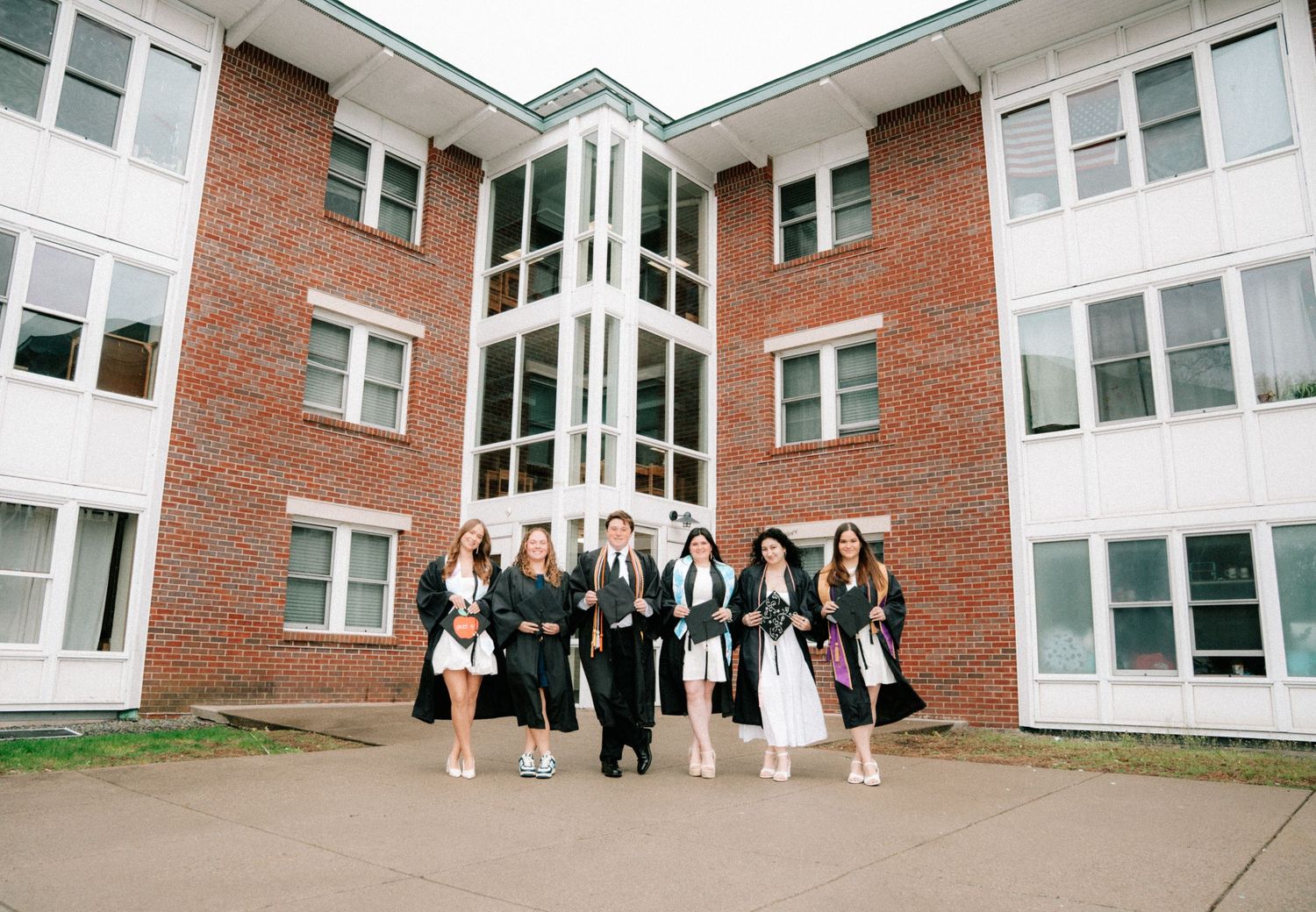 Group of graduates in black robes walking together in front of a brick university building.