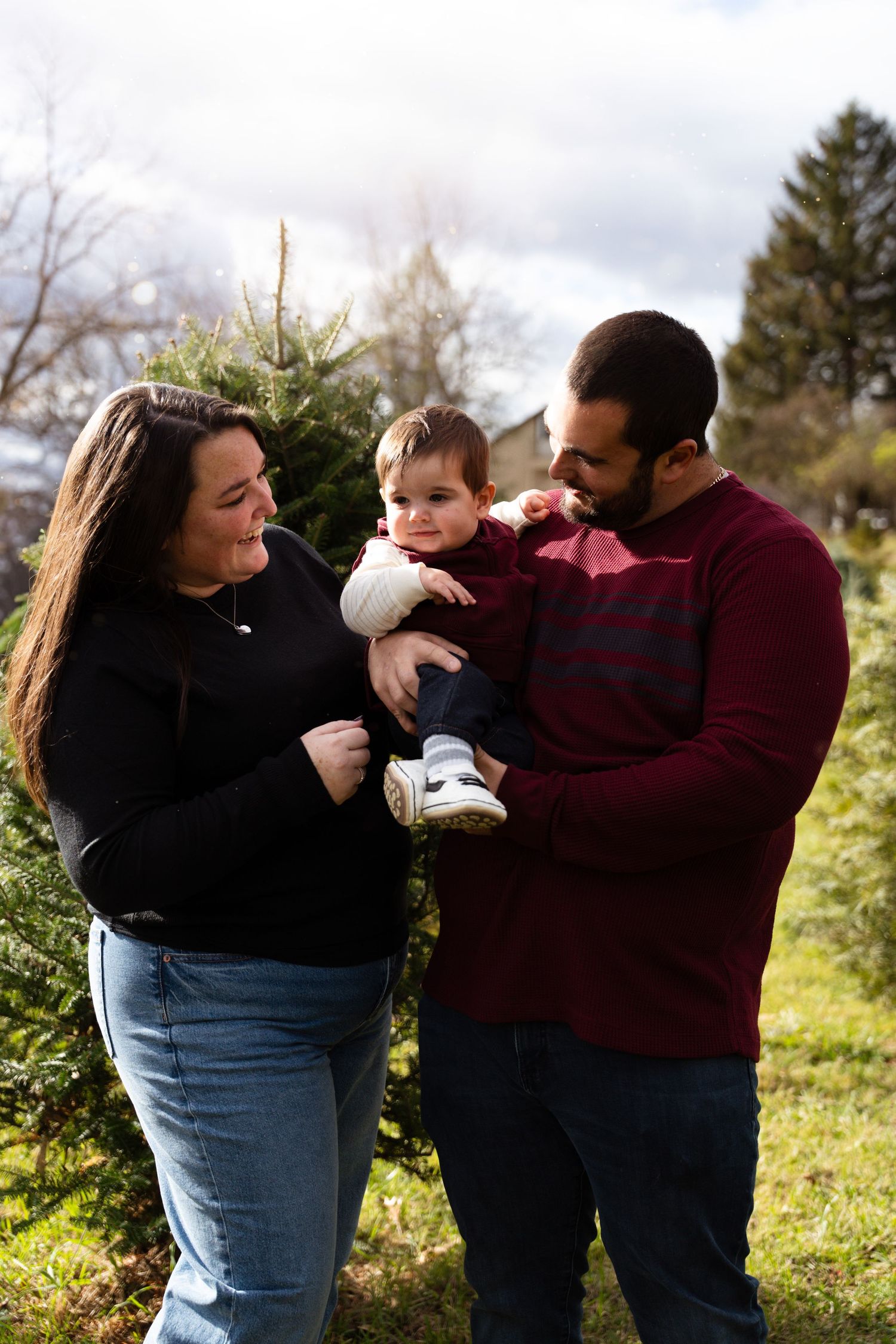 Happy family with mother, father, and baby standing outdoors near evergreen trees.