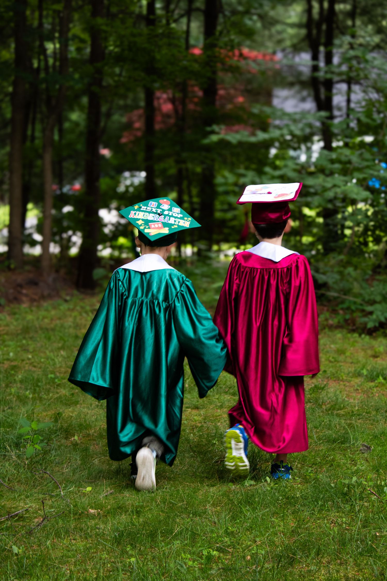 Two young graduates in green and red cap and gown walk hand in hand across a lawn.