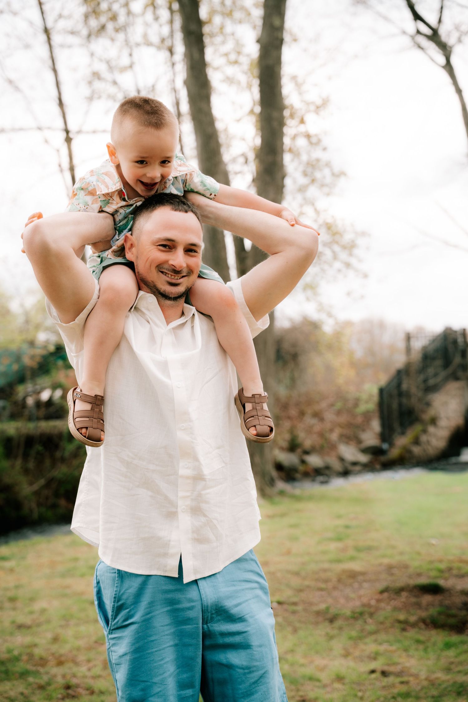 A smiling father carries his laughing young son on his shoulders outdoors in a green park setting.