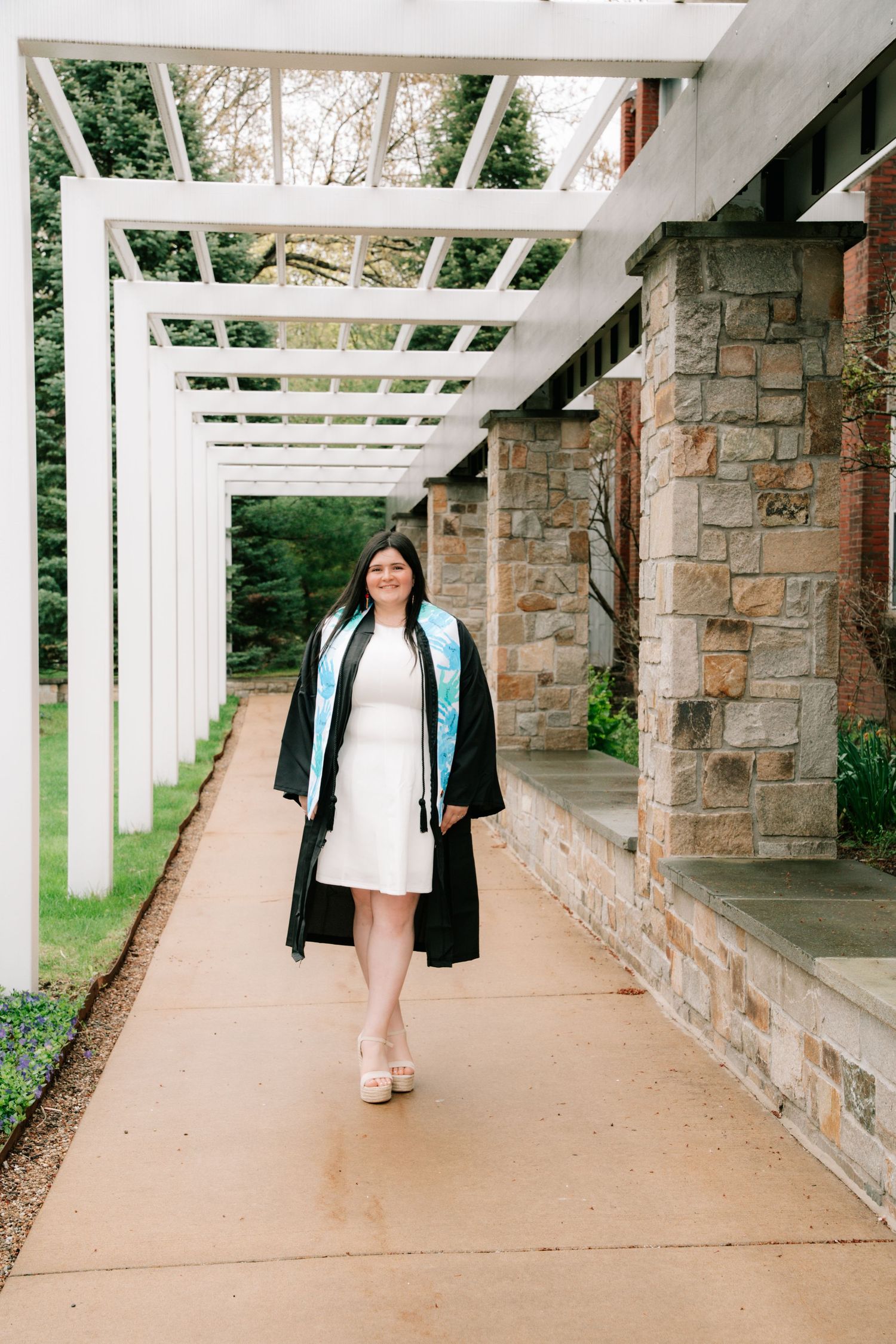 Female graduate in black robe with light blue stole walking under white pergola at university.