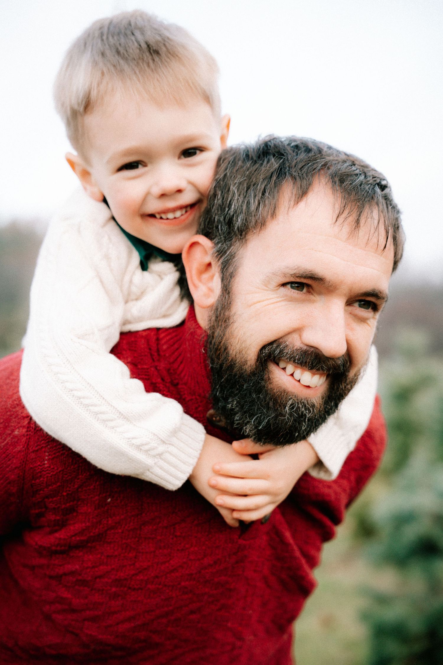 A bearded father smiles while giving his young blonde son a piggyback ride outdoors.