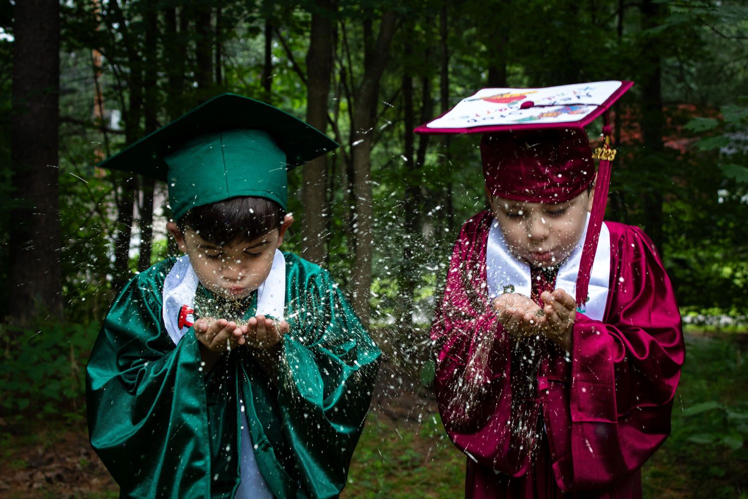 Two young children in graduation caps and gowns blowing glitter from their hands in a forest.