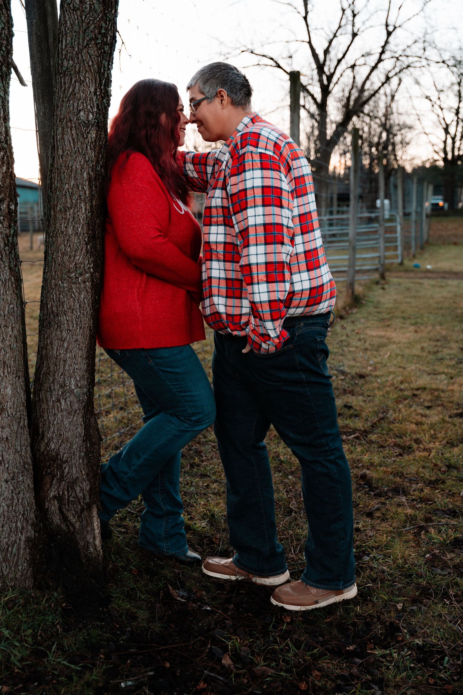 A romantic couple leans against a tree at dusk, touching foreheads in a tender moment outdoors.