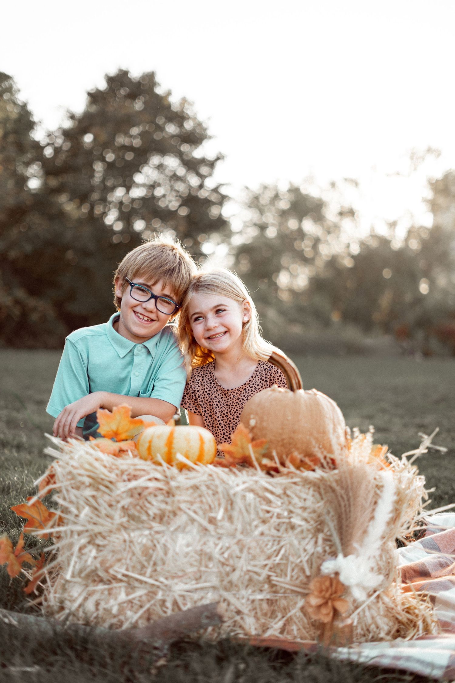 Two smiling children pose beside a hay bale with pumpkins and autumn leaves outdoors during golden hour.