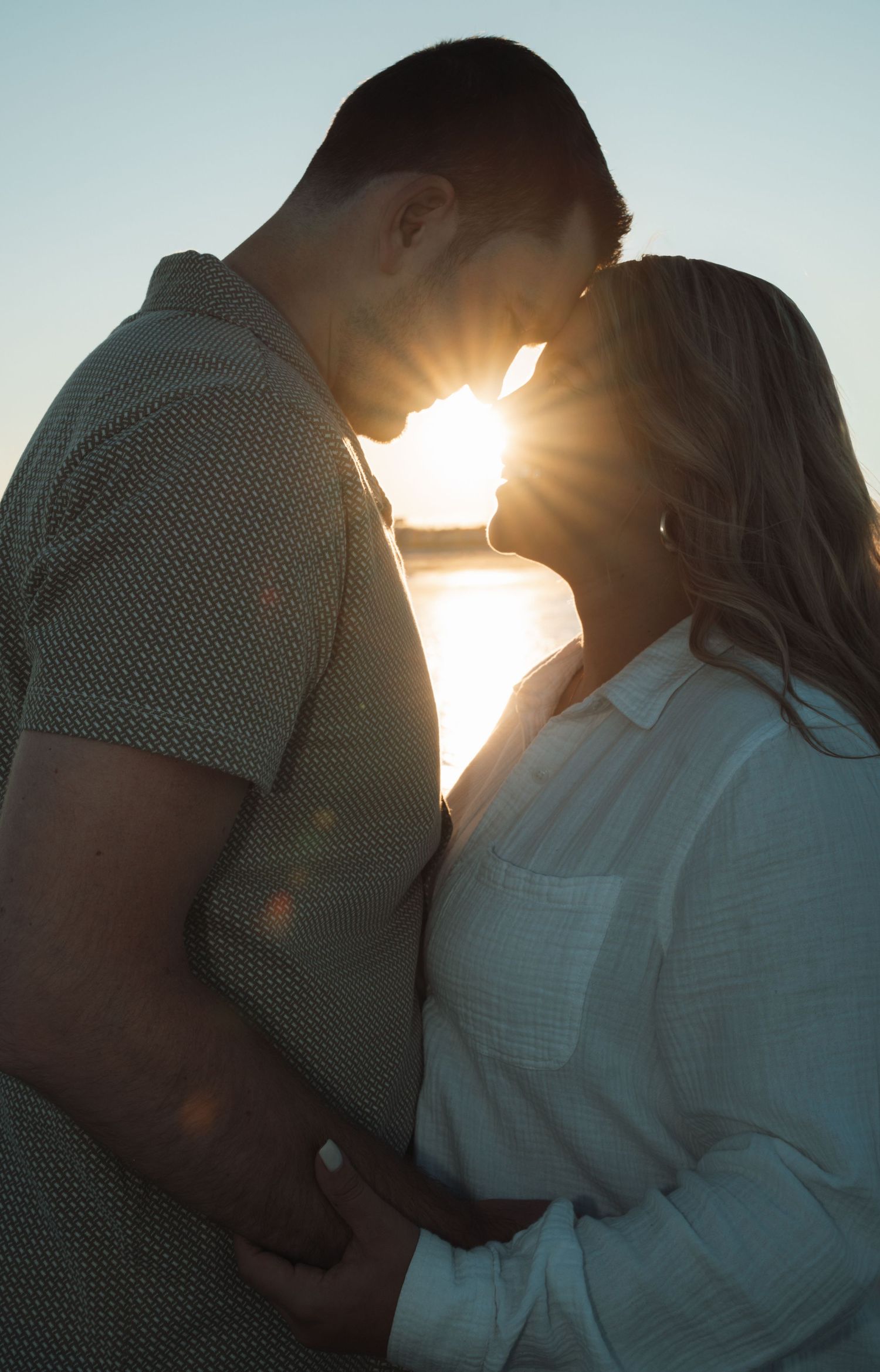 Couple sharing a romantic sunset kiss silhouetted against golden sun flare near water.
