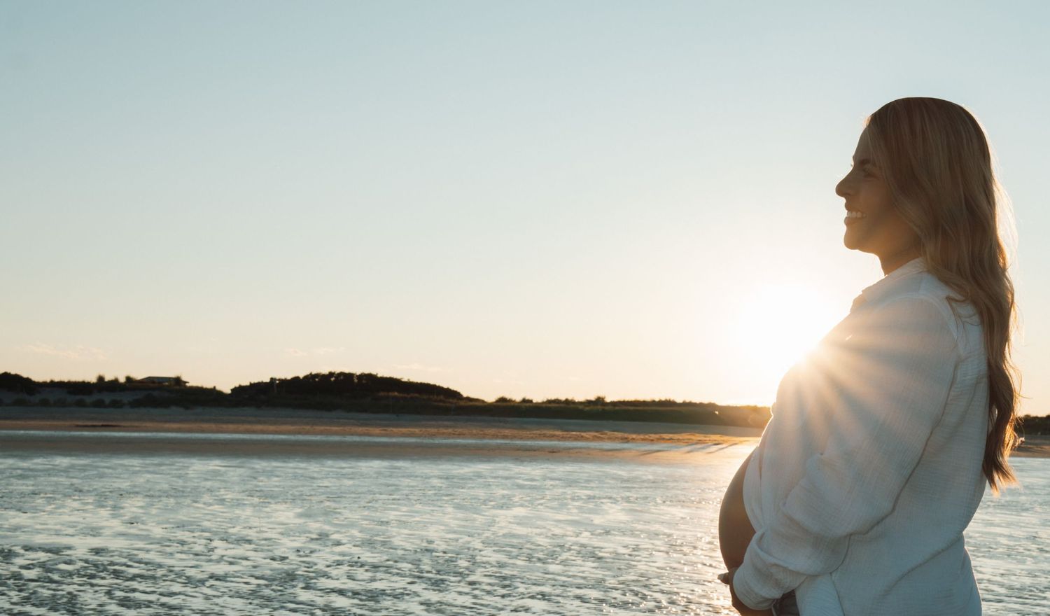 Pregnant woman in white shirt standing on sunny beach at golden hour, cradling her baby bump by the ocean.