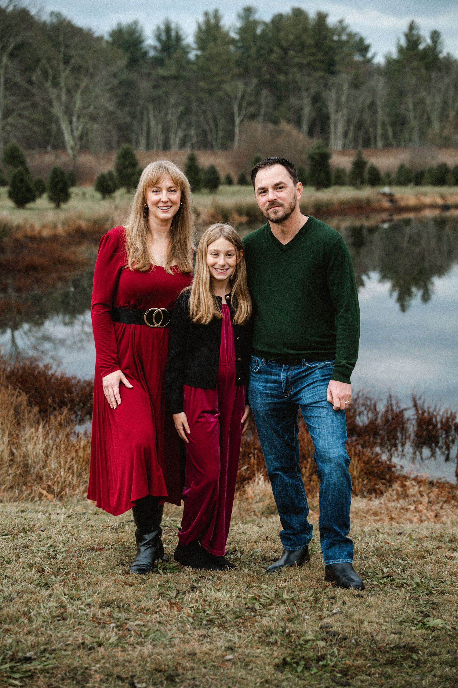 A family of three poses together near a reflective pond at a Christmas tree farm in festive attire.