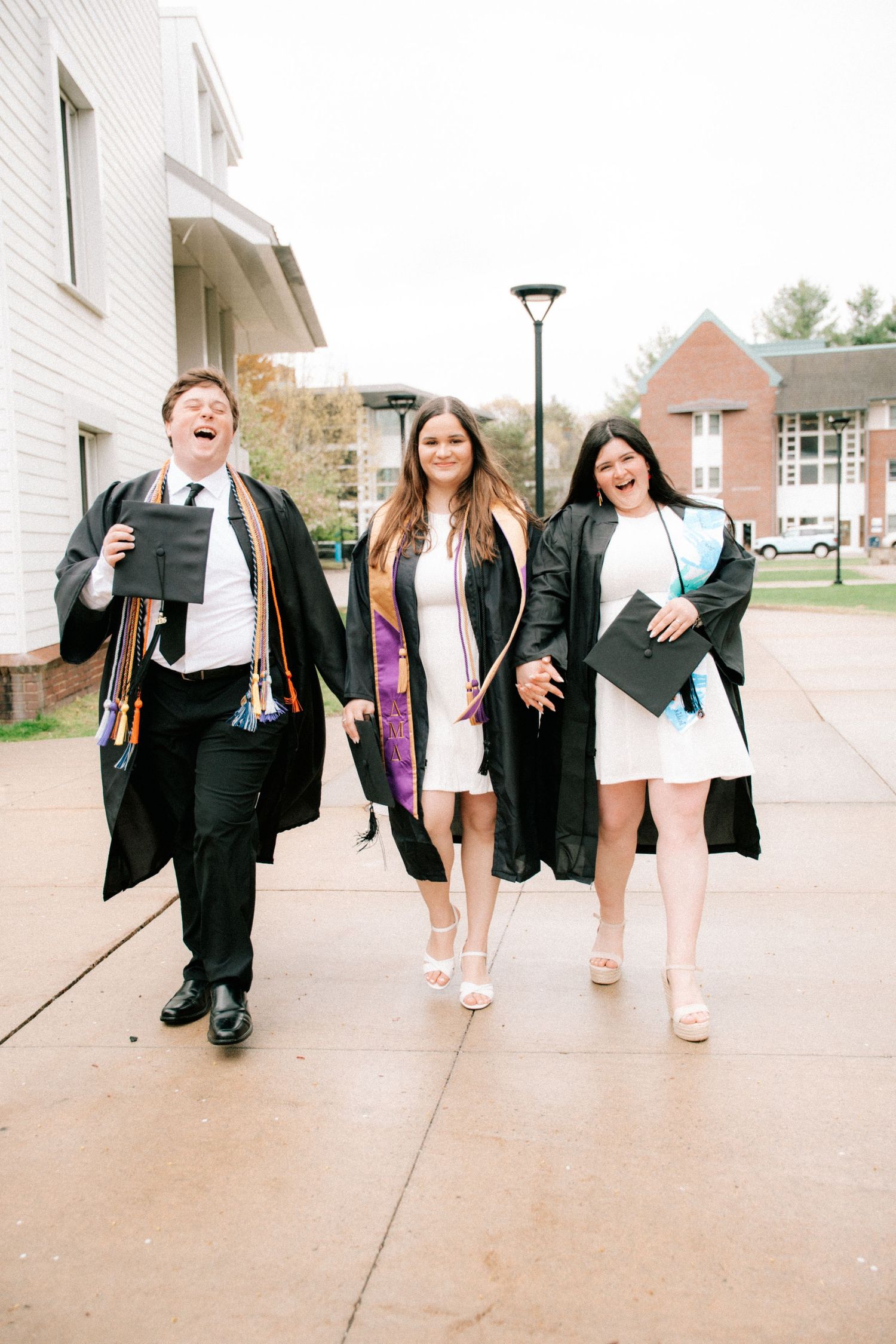 Three graduates in black gowns walk and laugh together on a college campus pathway.