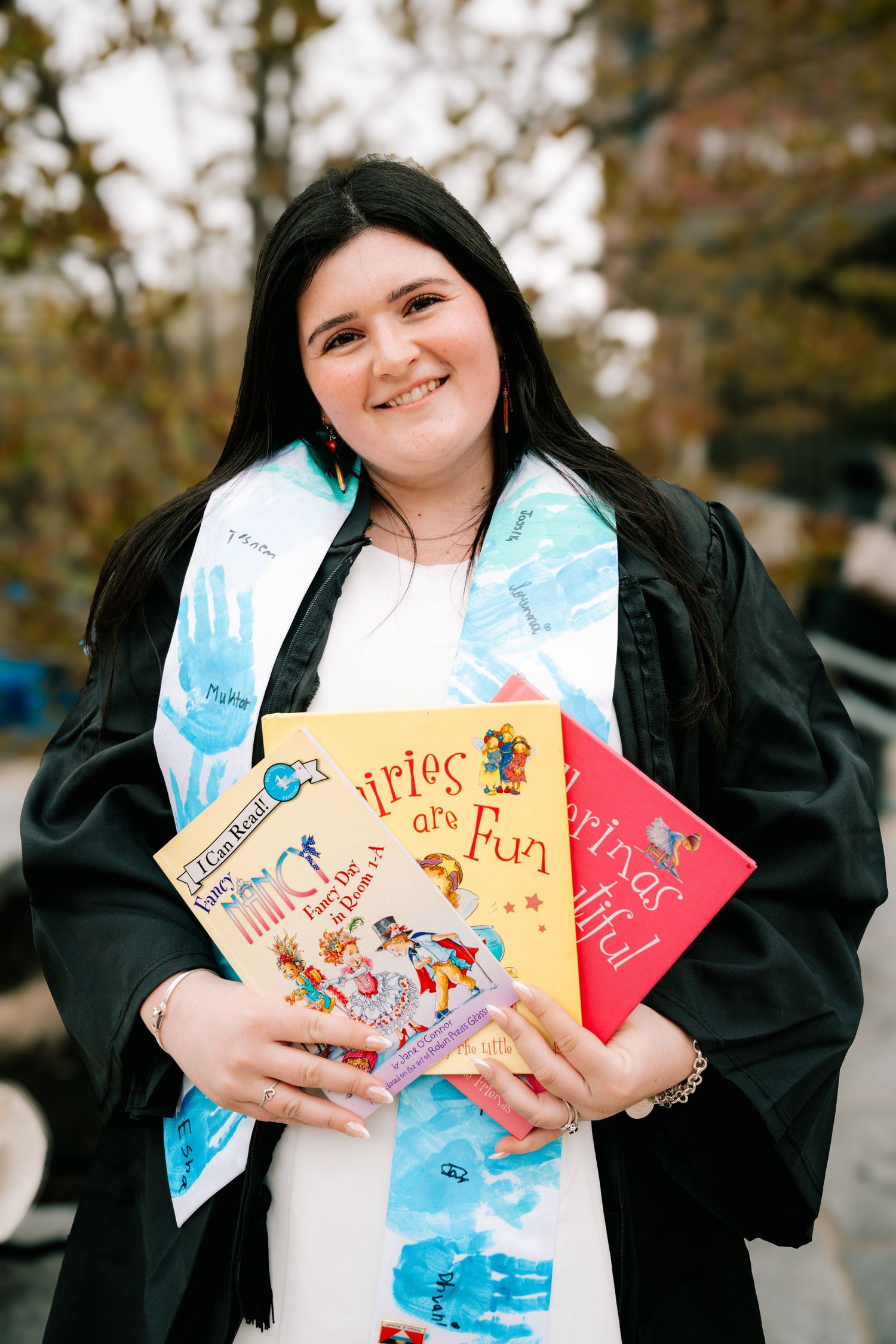 A happy graduate in black gown holds colorful children's books while wearing a handprint-decorated stole.