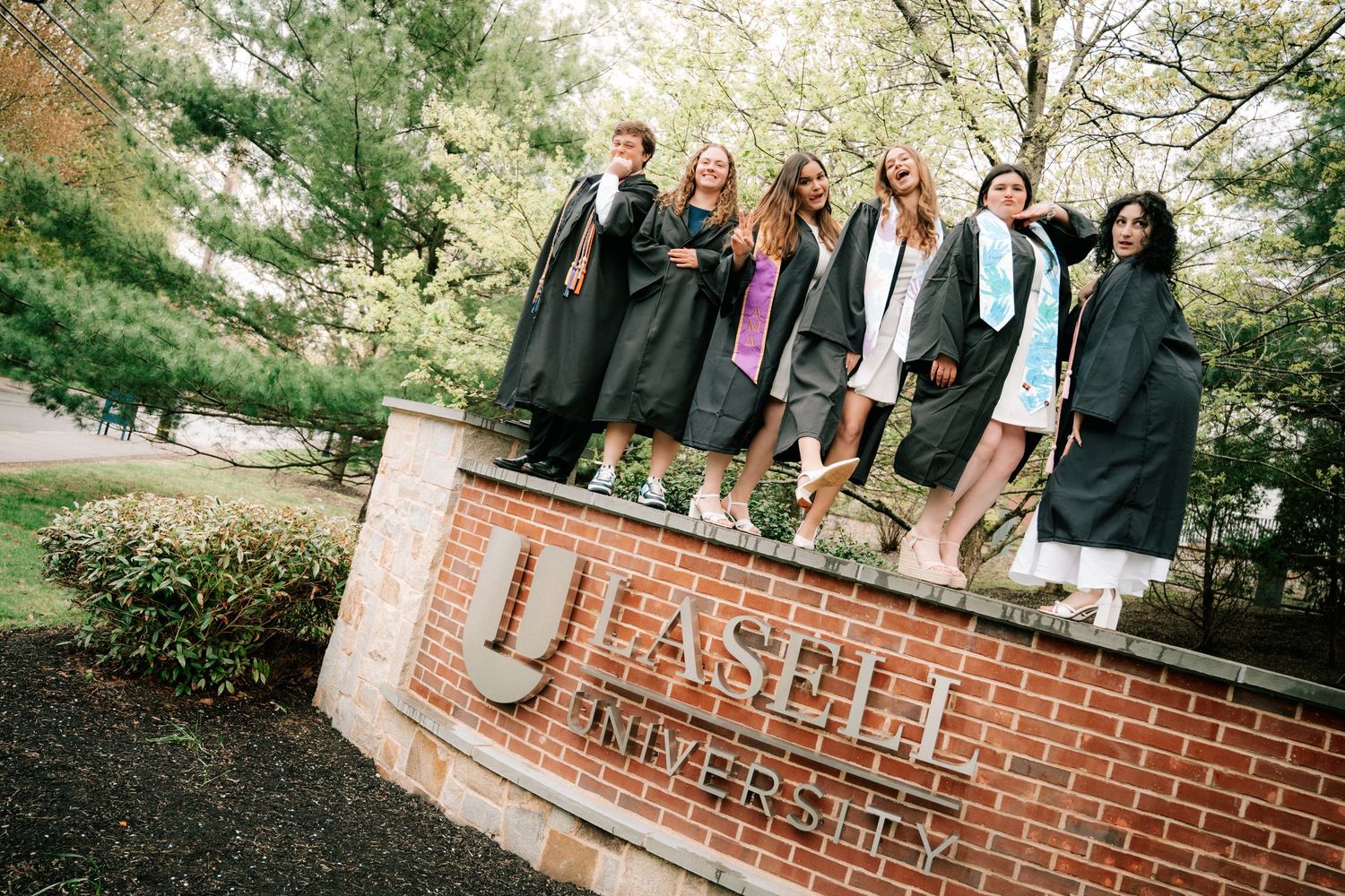 Group of graduates in black robes standing on Lasell University brick entrance sign outdoors.