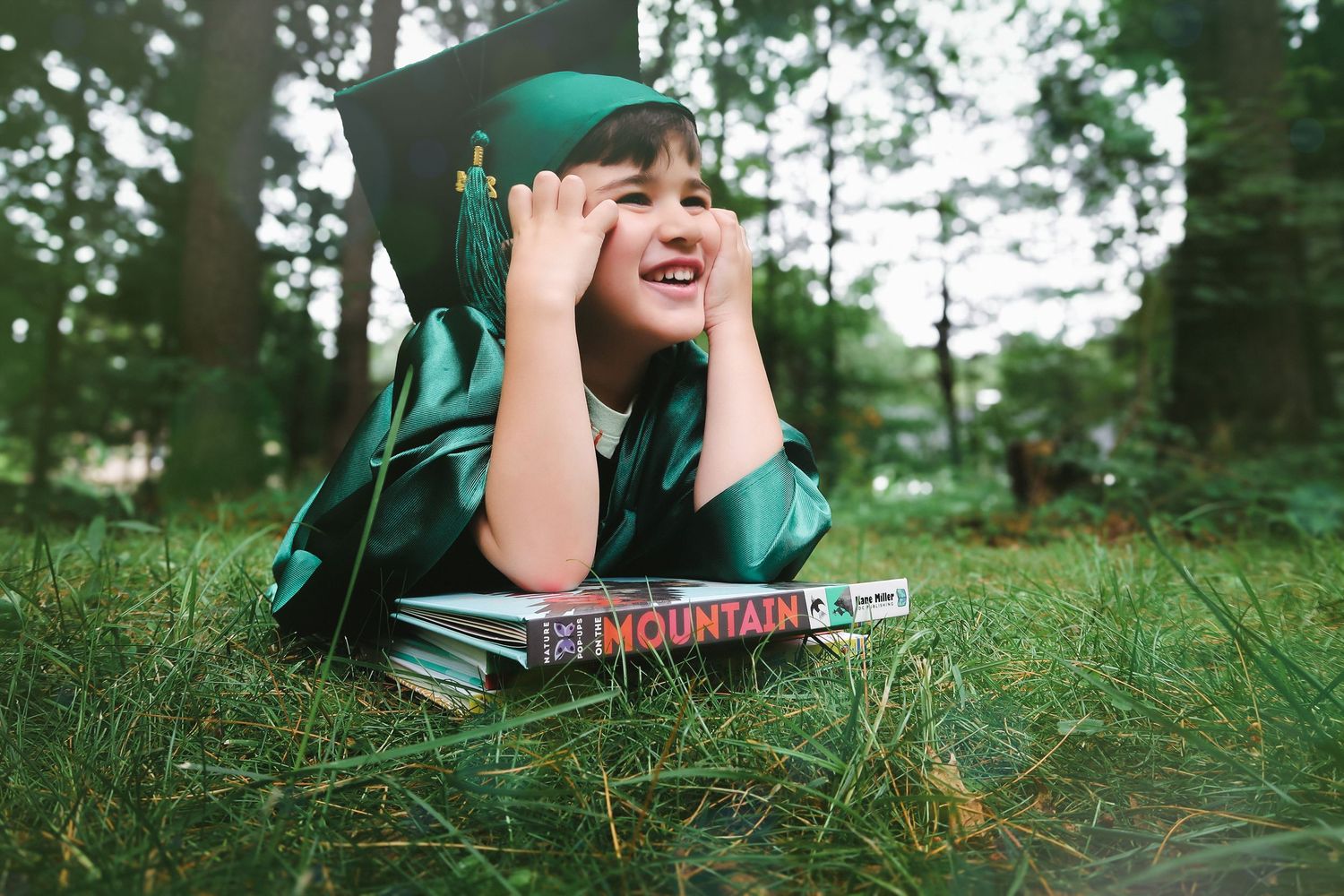 A smiling child in a green graduation cap and gown lies on grass resting on books.
