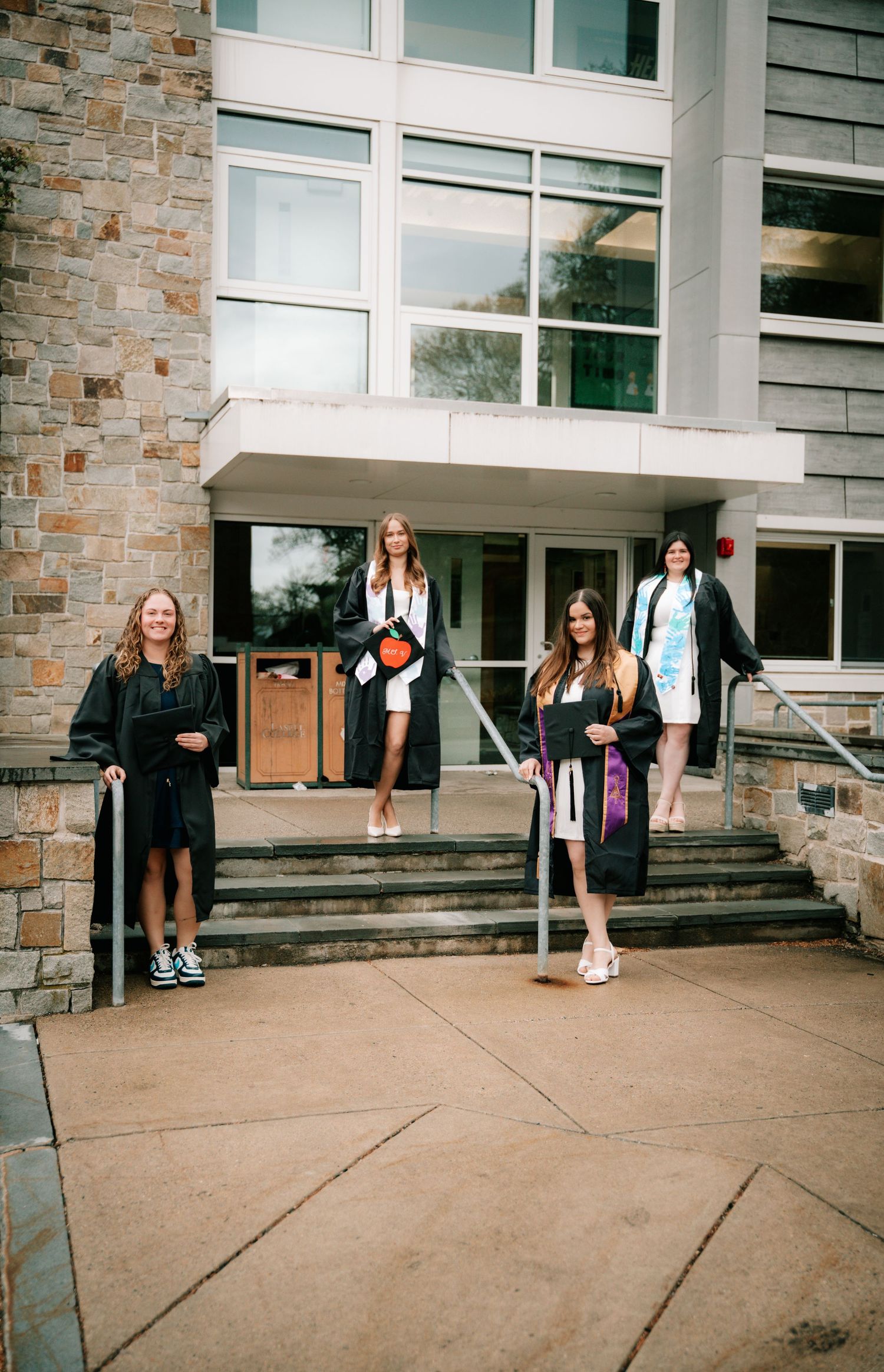 Four graduates in black robes posing on steps outside a modern stone university building.