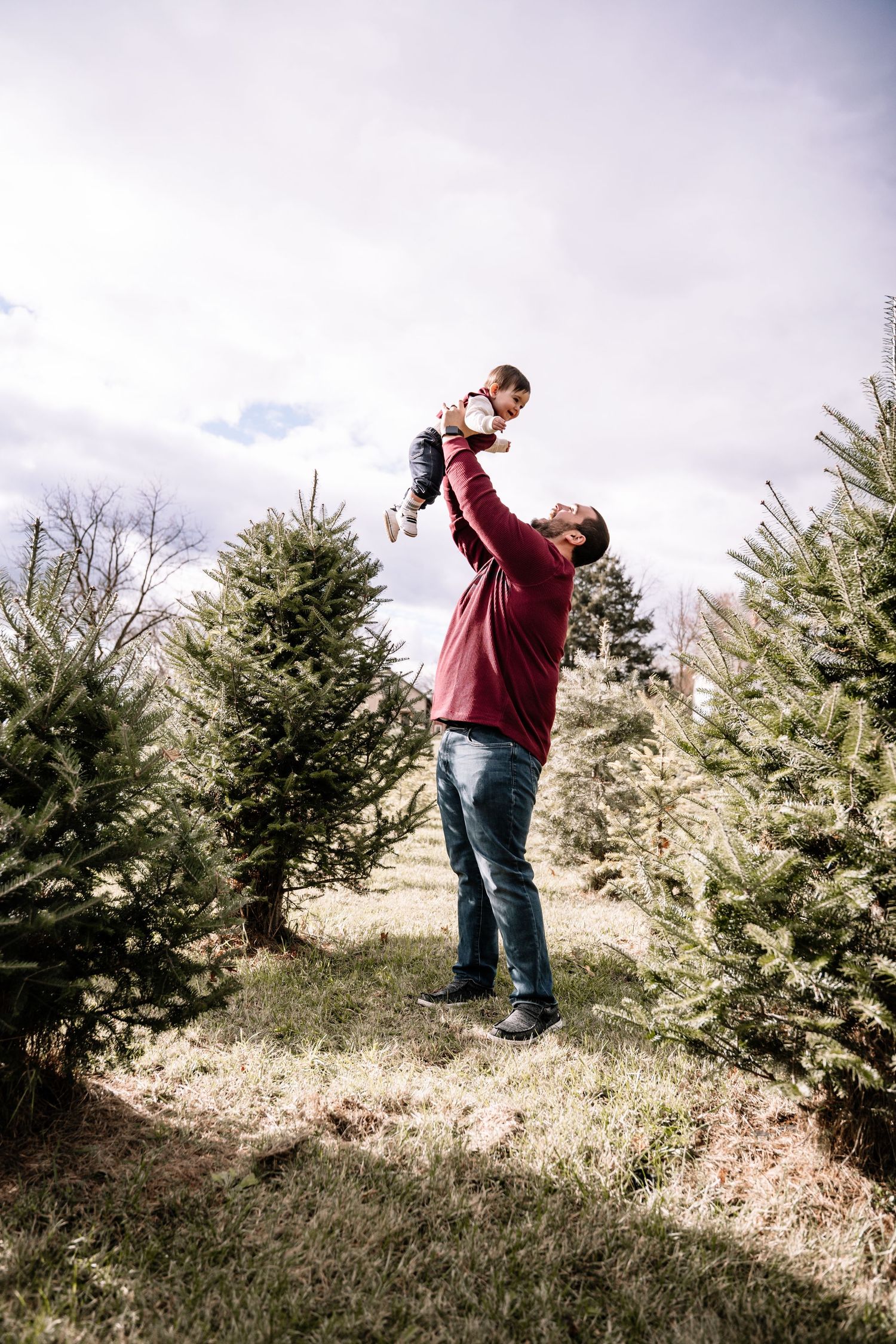 Father lifting baby high in the air surrounded by Christmas trees on a farm.