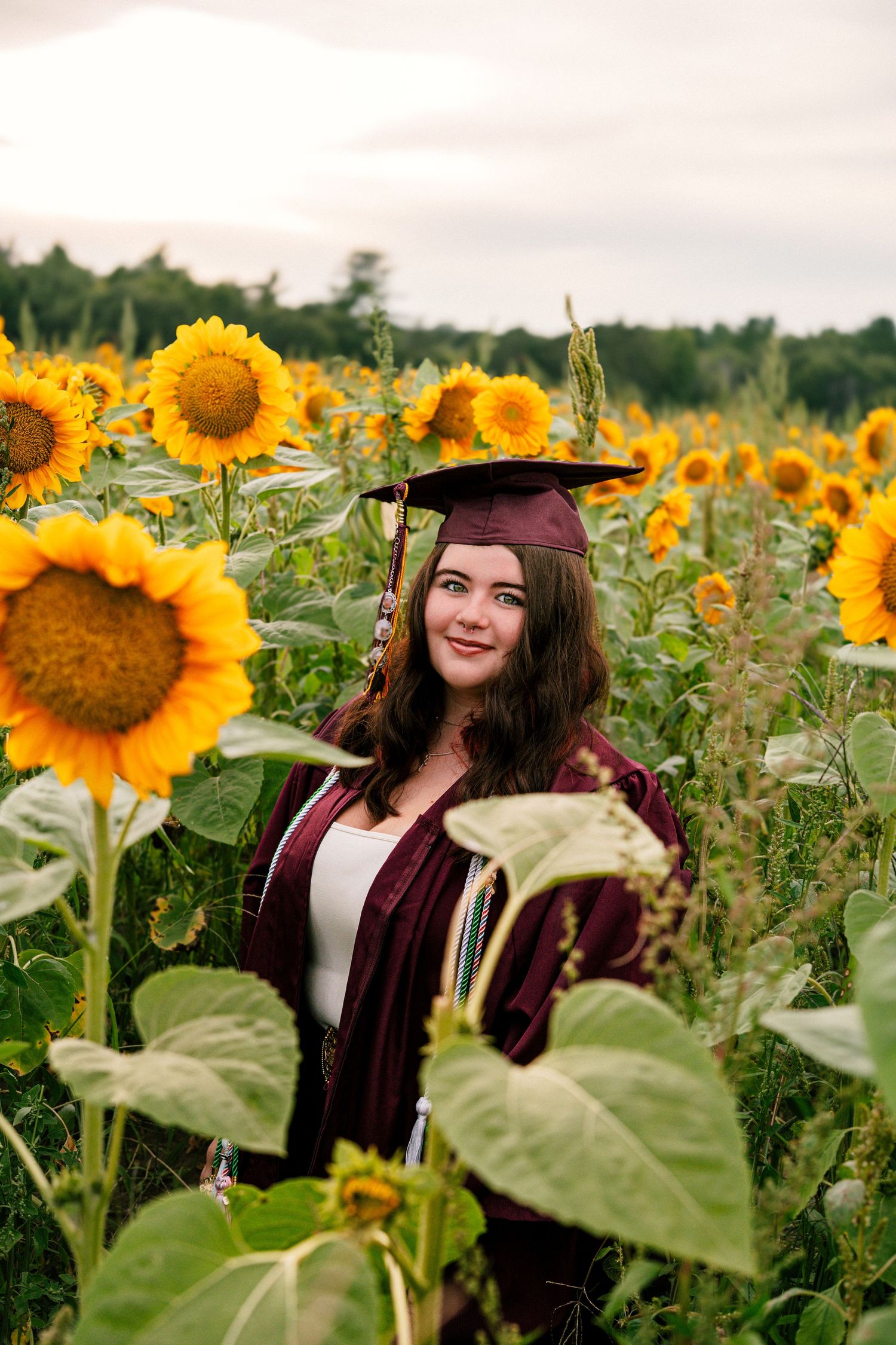 Young woman in maroon graduation cap and gown smiling in a vibrant sunflower field at sunset.