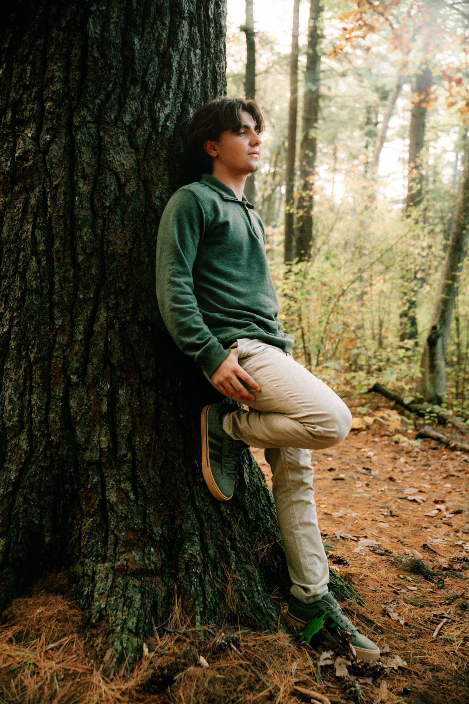 Teen boy in green hoodie leaning against a large tree in an autumn forest.