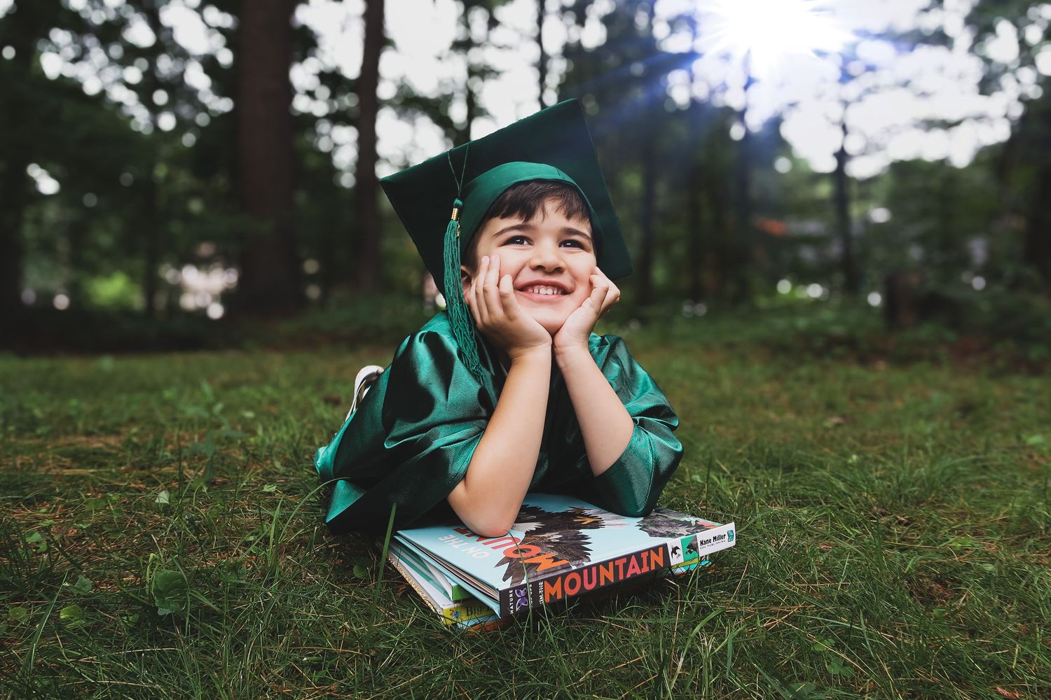 A young child in a green graduation cap and gown lies on grass smiling with books beneath.
