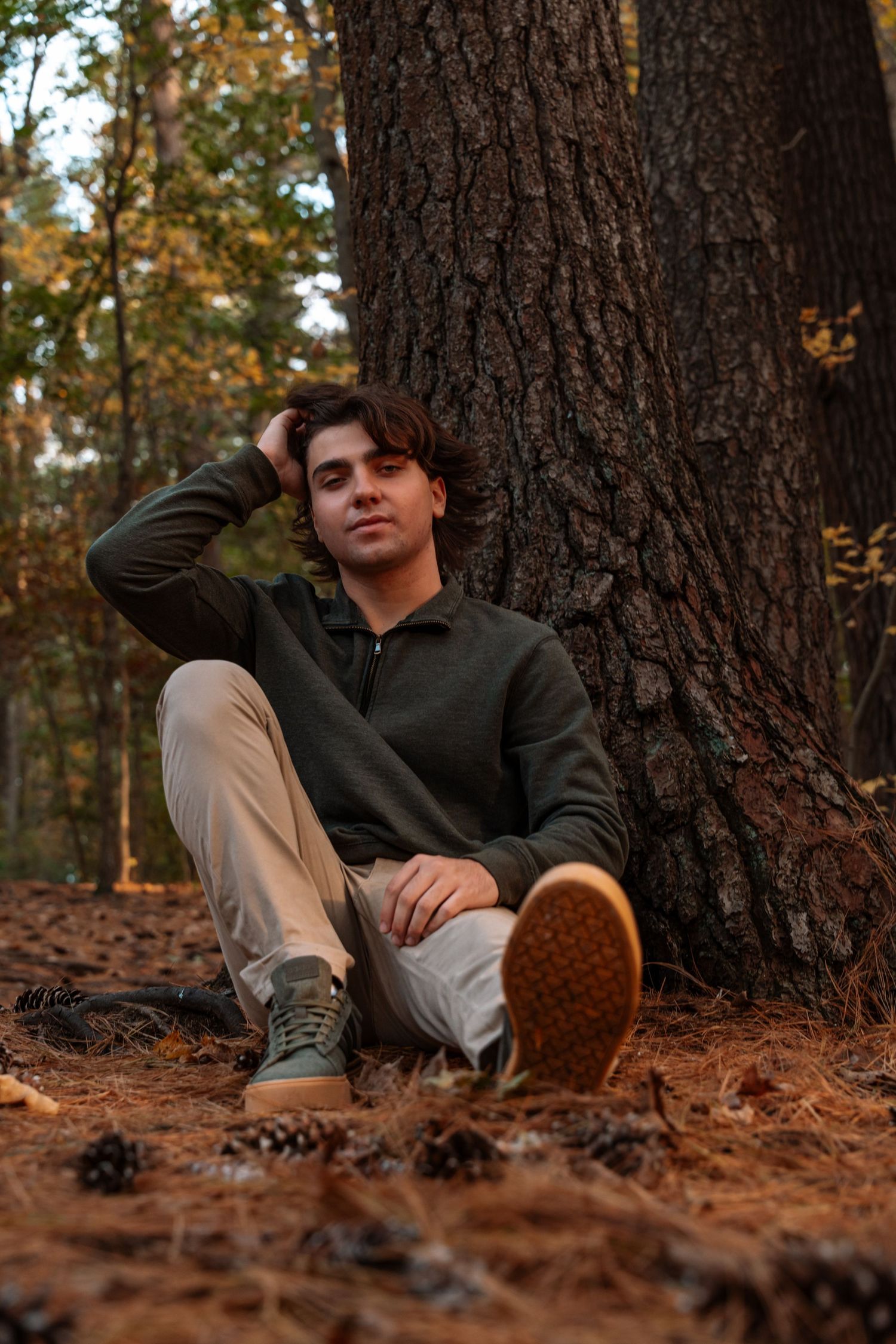 Young man in green hoodie sits against pine tree with pinecones on forest floor.