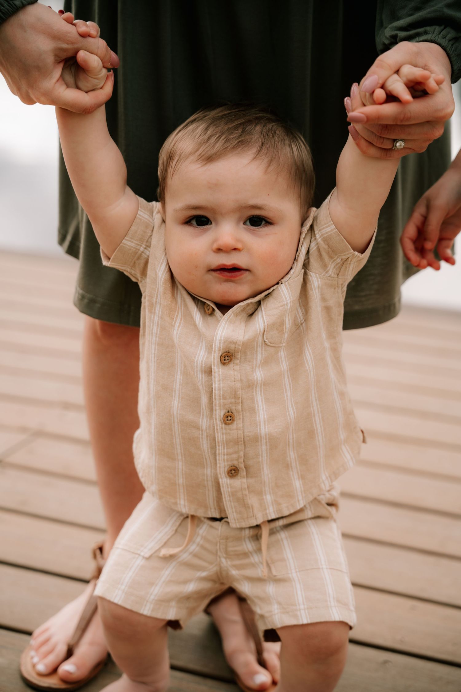Adorable baby boy in striped linen outfit standing on a wooden deck held by parent hands.