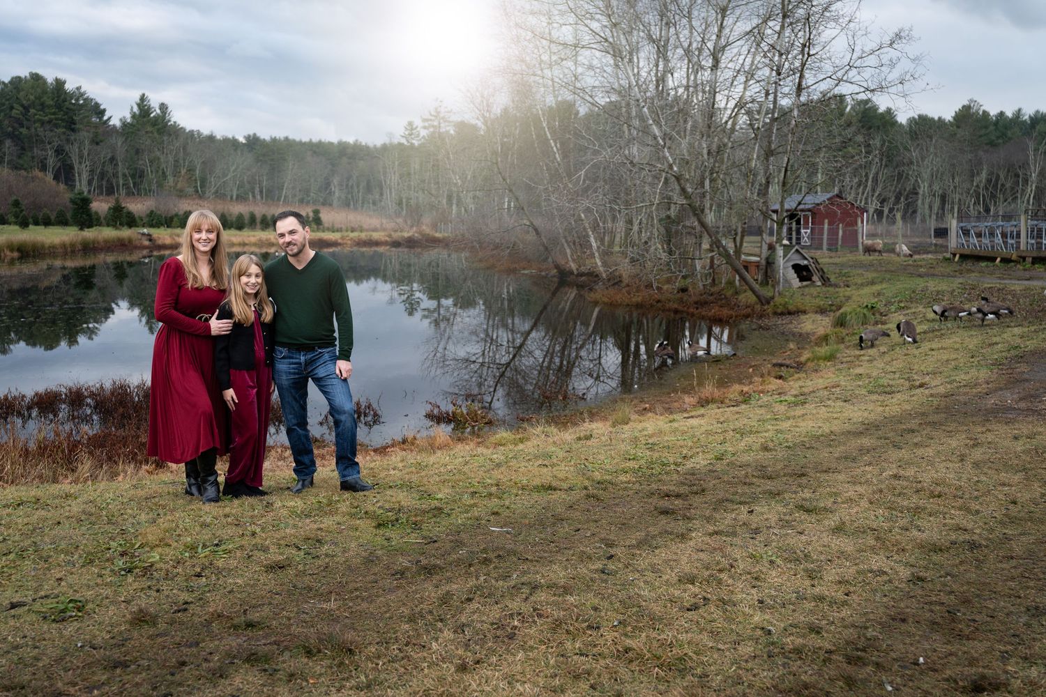 Family of three posing by a scenic pond with a red barn in the background.