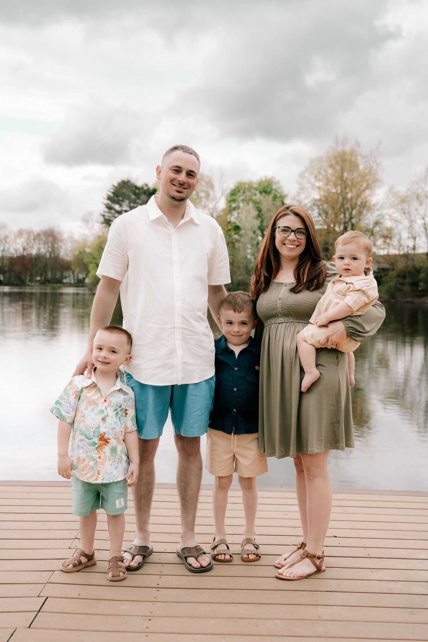 Happy family of five posing on a wooden dock by a lake on a cloudy spring day.