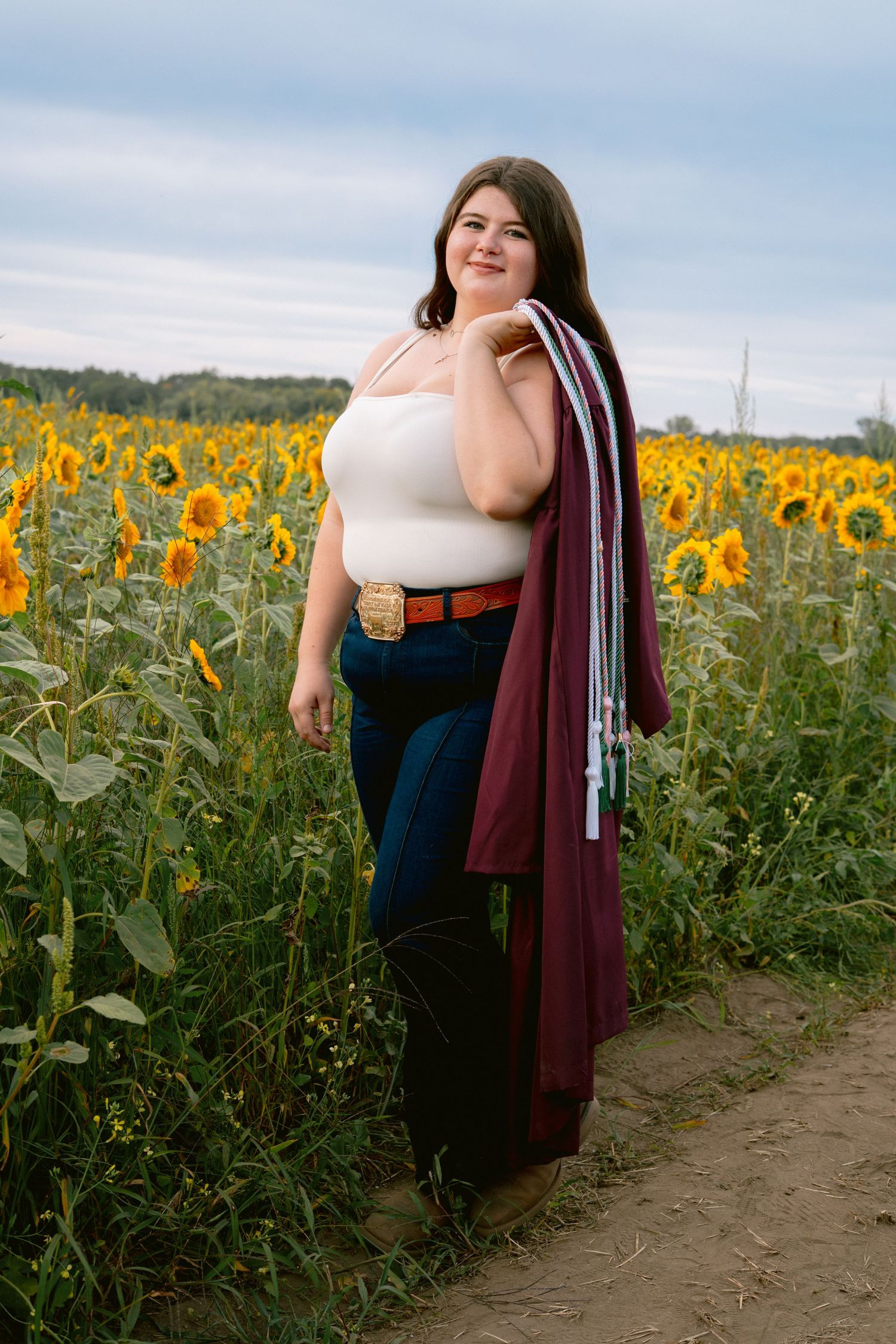 Graduate in maroon gown holds regalia over shoulder in a sunflower field.