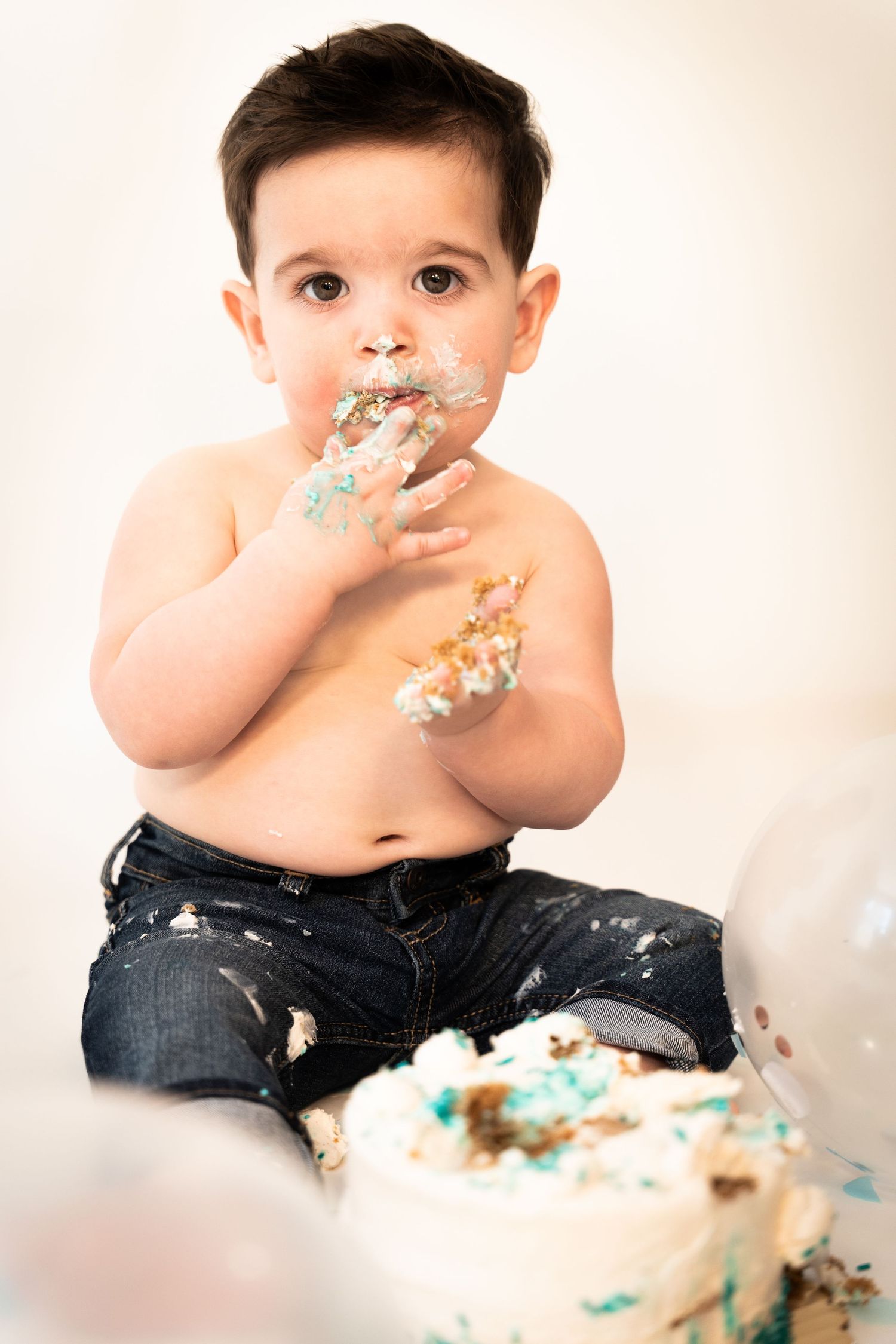 An adorable baby boy enjoys his first birthday cake smash, covered in frosting during a studio photo session.