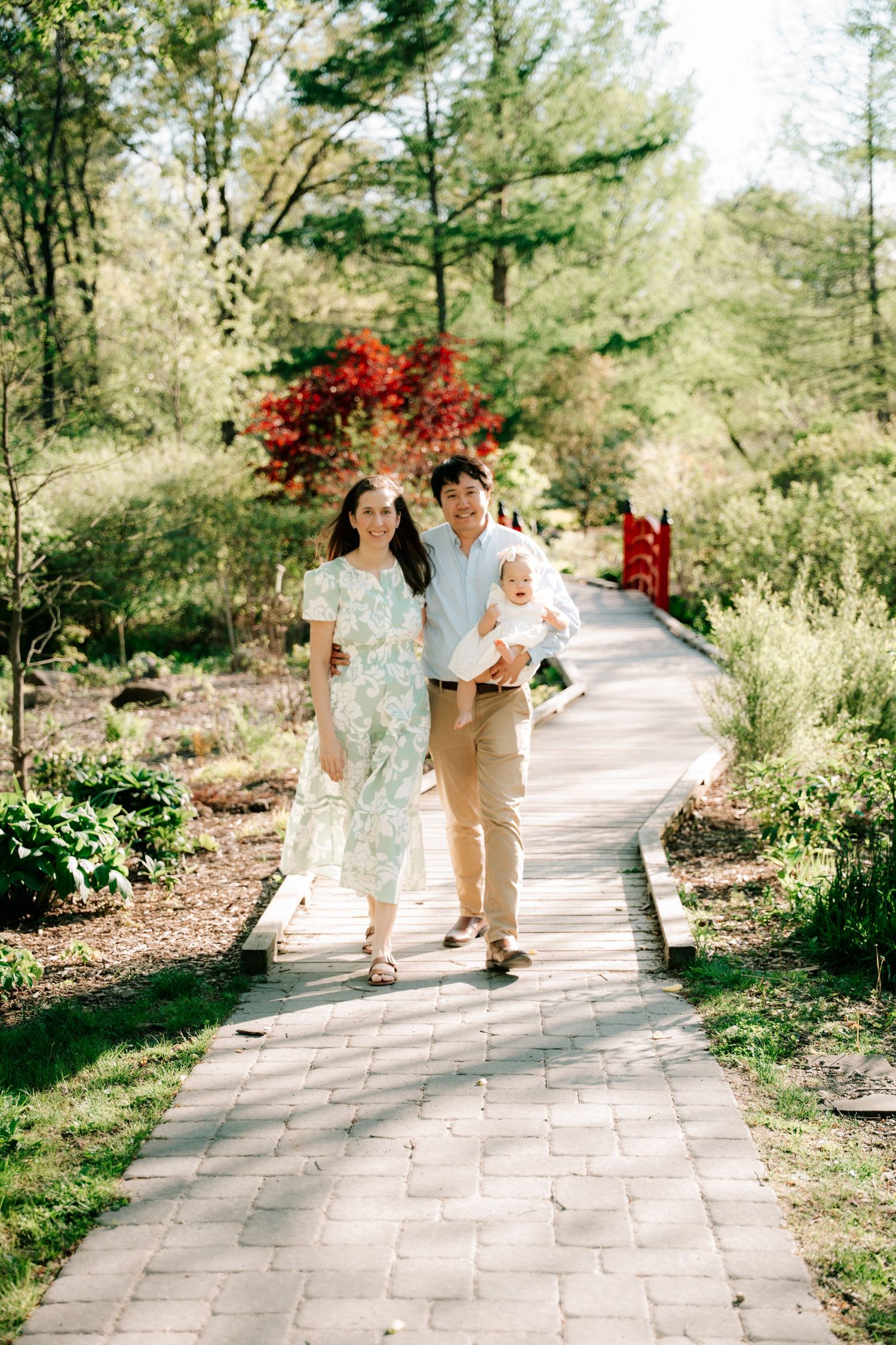 A happy family of three walks along a garden path, with lush greenery and a red bridge in the background.