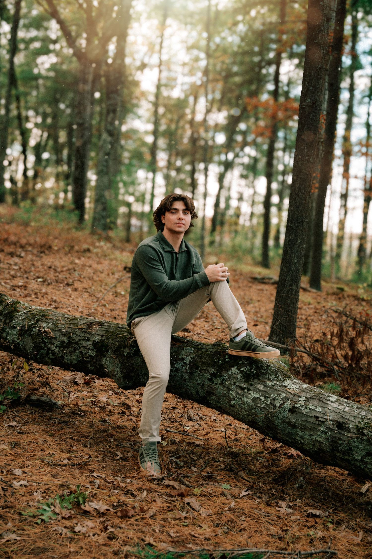 Young man in green hoodie sits on fallen log in autumn forest with sunlight.