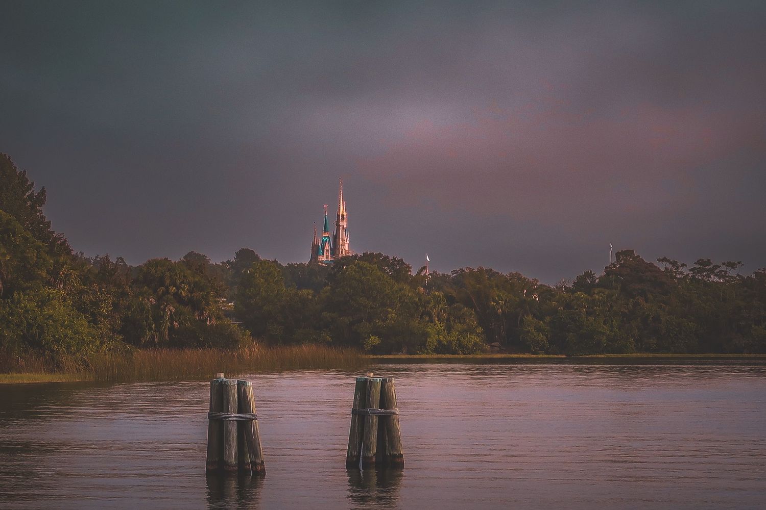 Distant fairytale castle spires rise above lush green trees reflecting in calm river under dramatic cloudy sky.