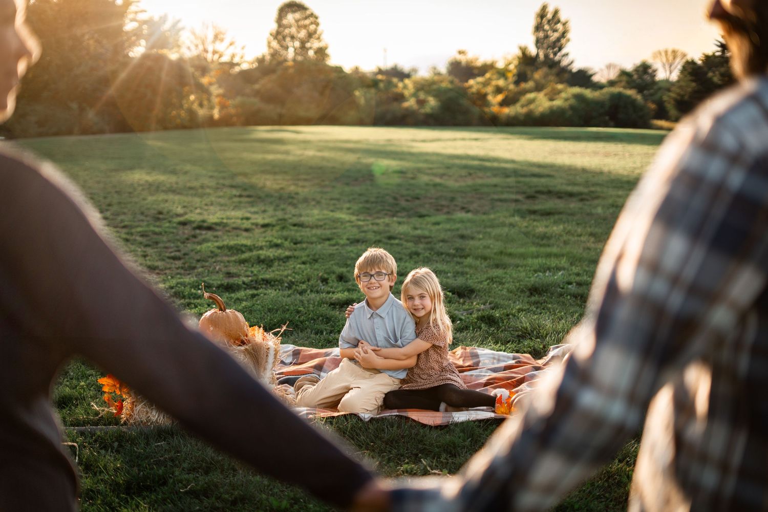 Two children sit on a picnic blanket in a sunny park while their parents hold hands in the foreground.