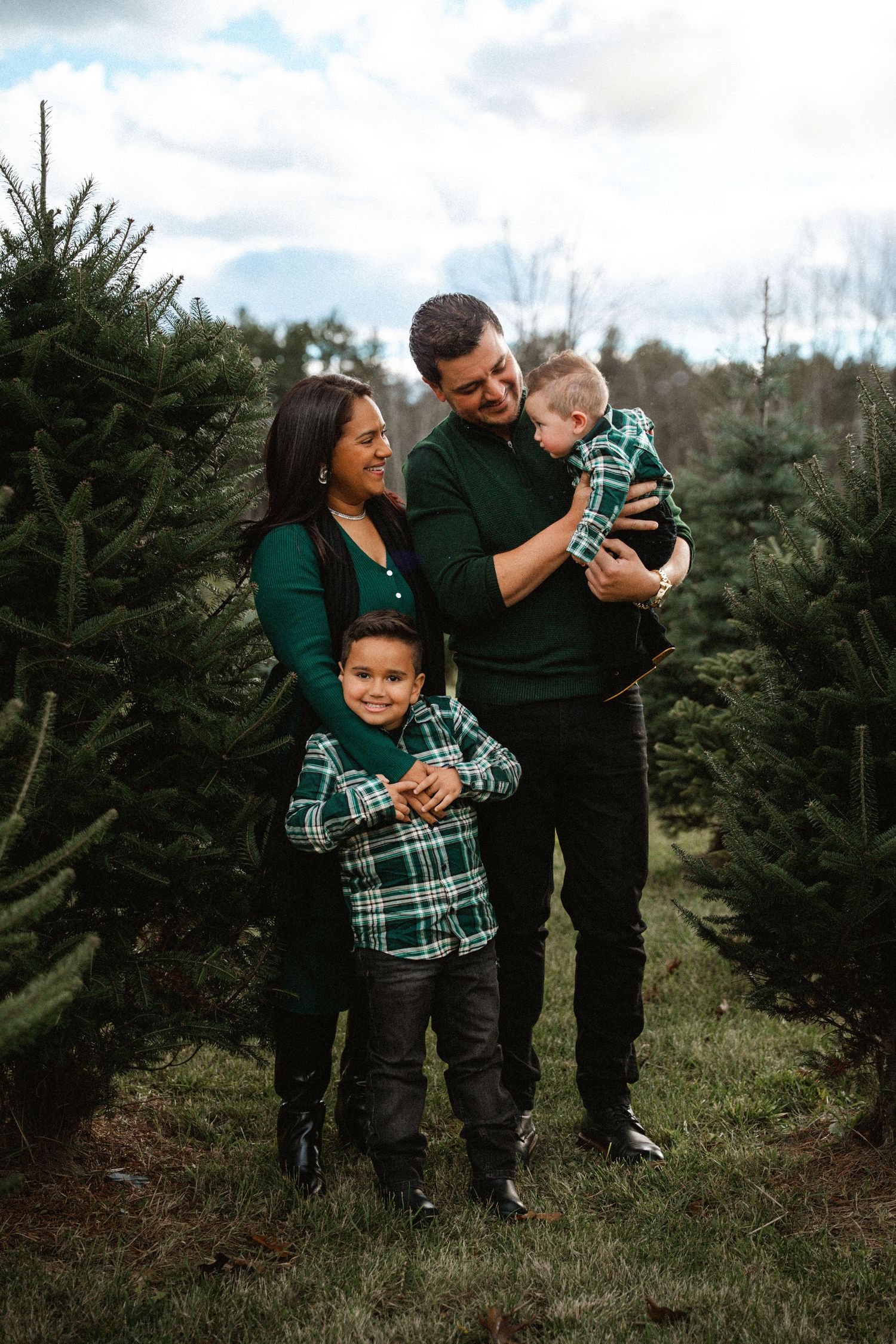 Happy family of four in green plaid outfits posing at a Christmas tree farm.