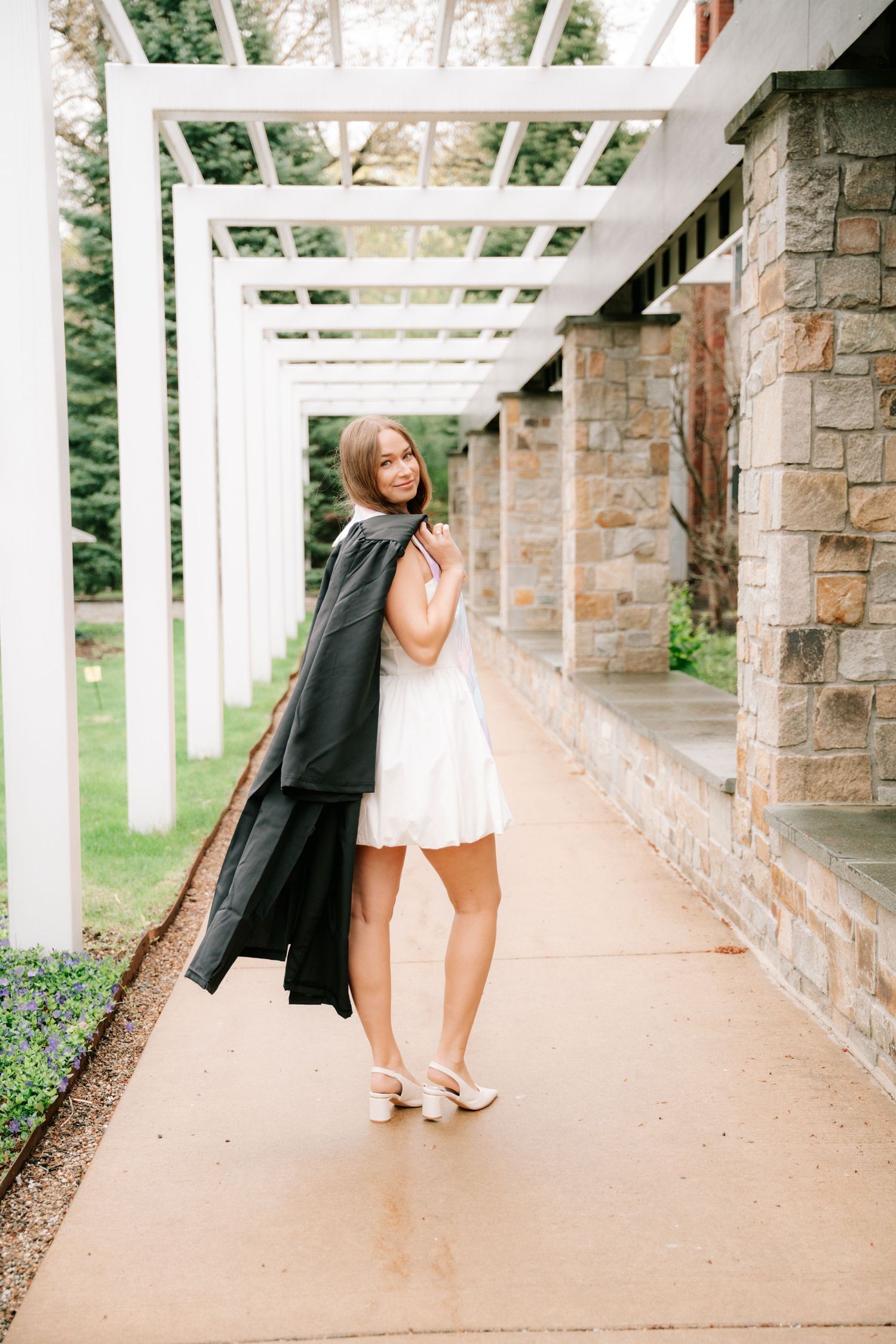 Female graduate in white dress holding black graduation robe while posing under white pergola.