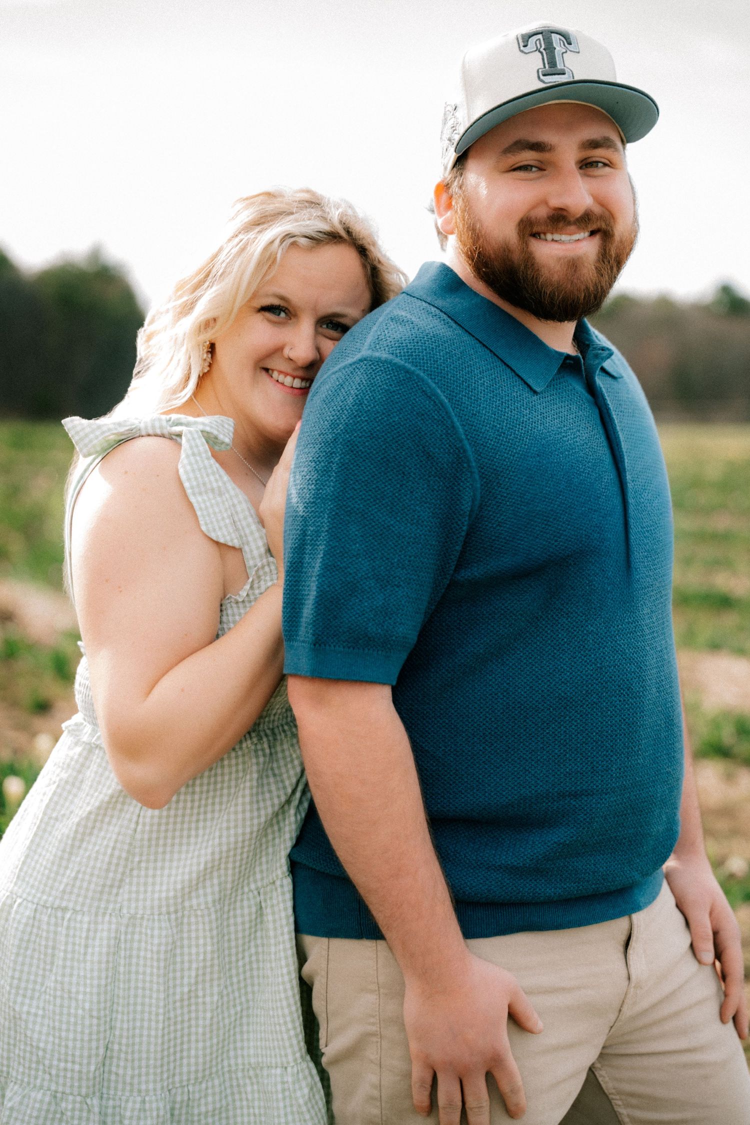 Smiling couple posing back-to-back in a field, man wearing a blue polo and baseball cap.