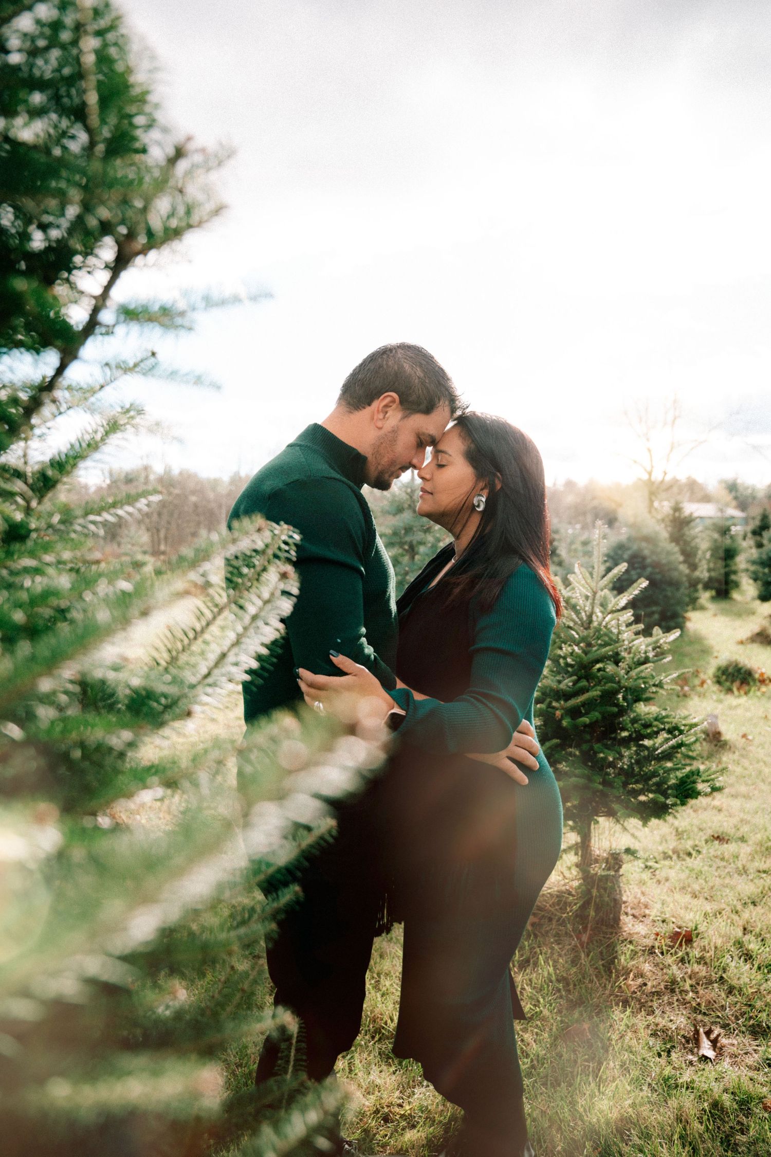 Couple embracing romantically among green Christmas trees at a tree farm, wearing matching dark green outfits.