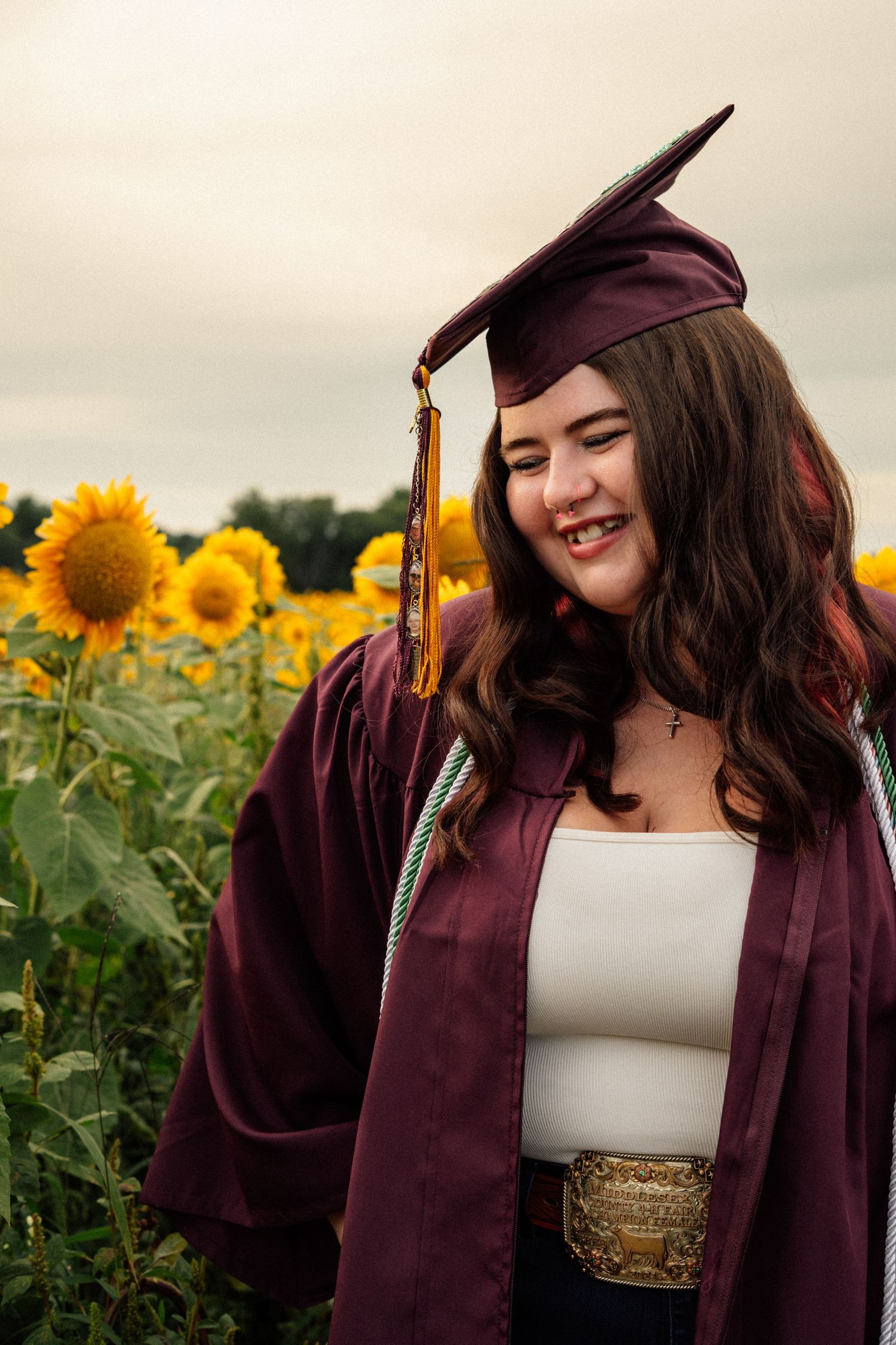 A laughing graduate in a maroon cap and gown poses among tall sunflowers in an outdoor field.