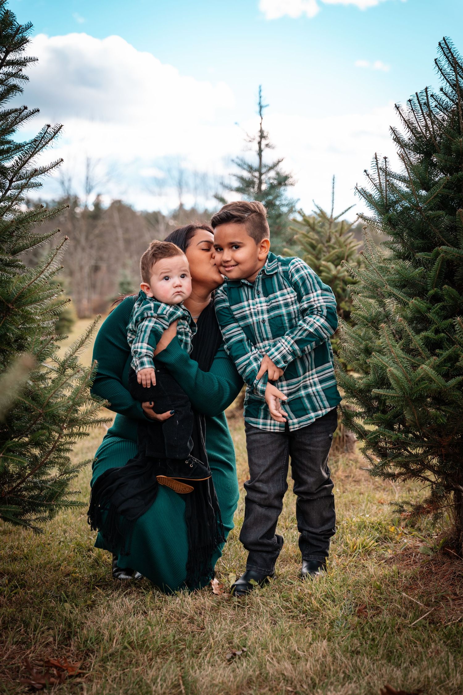 A mother kneels between her two young sons among Christmas trees, kissing her older boy on the cheek.