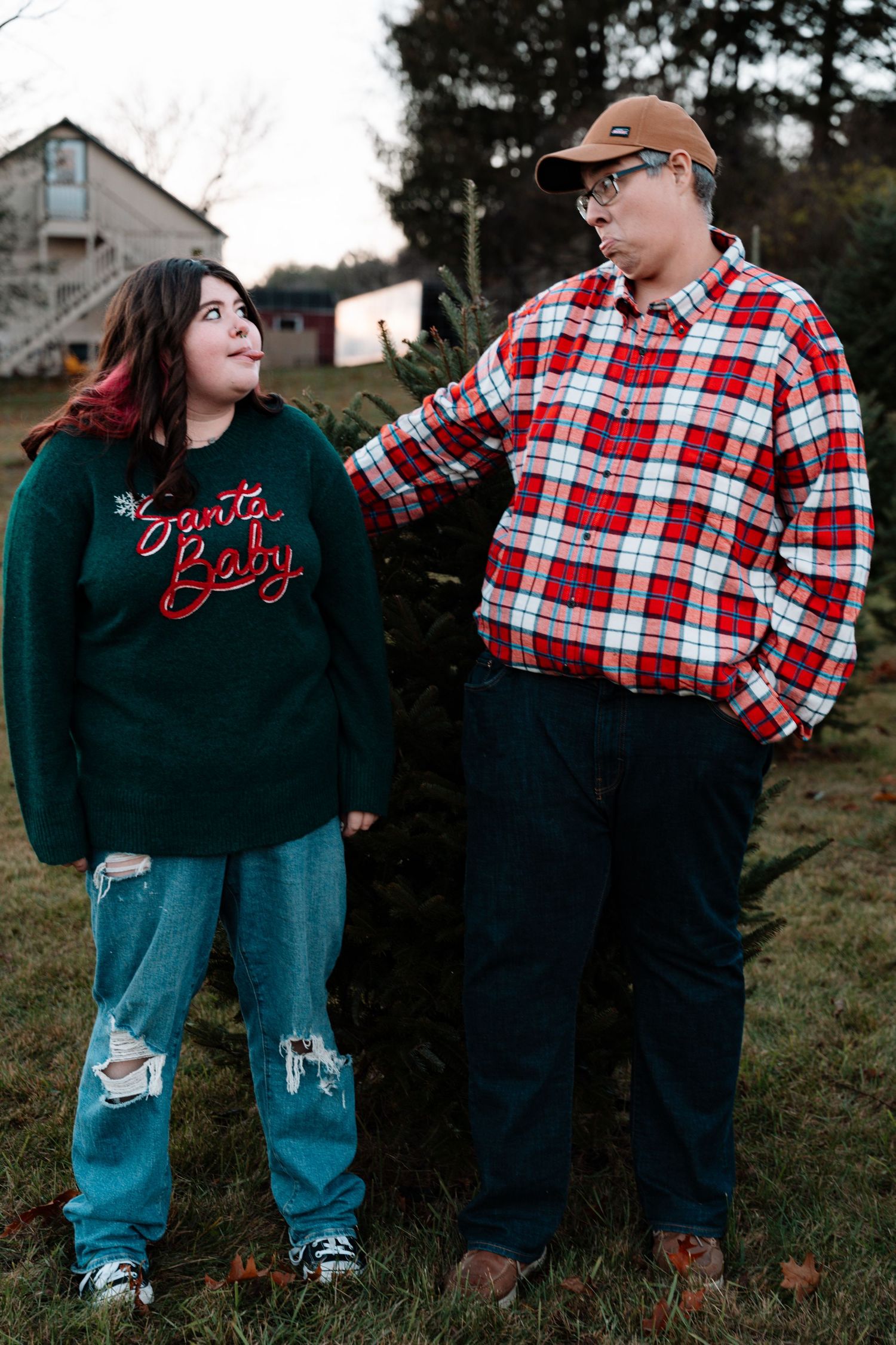 A teenage girl and boy stand together near a Christmas tree on a farm during a holiday photo session.