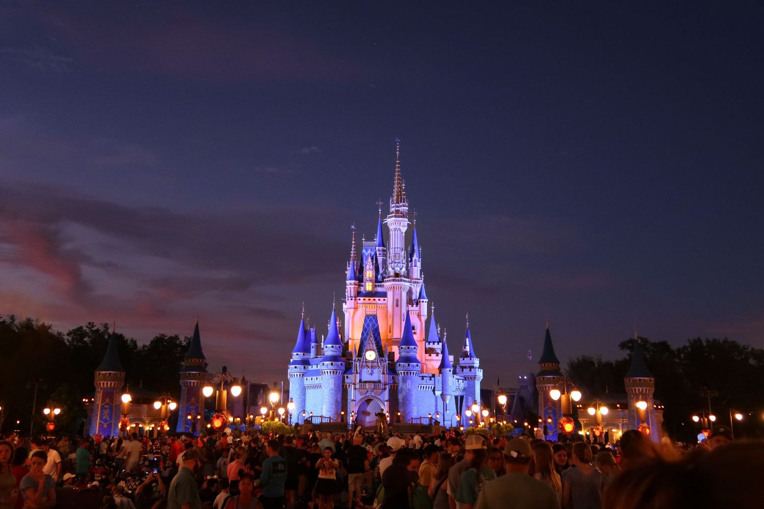 Cinderella Castle at Walt Disney World illuminated in purple and blue lights at dusk with crowds below.