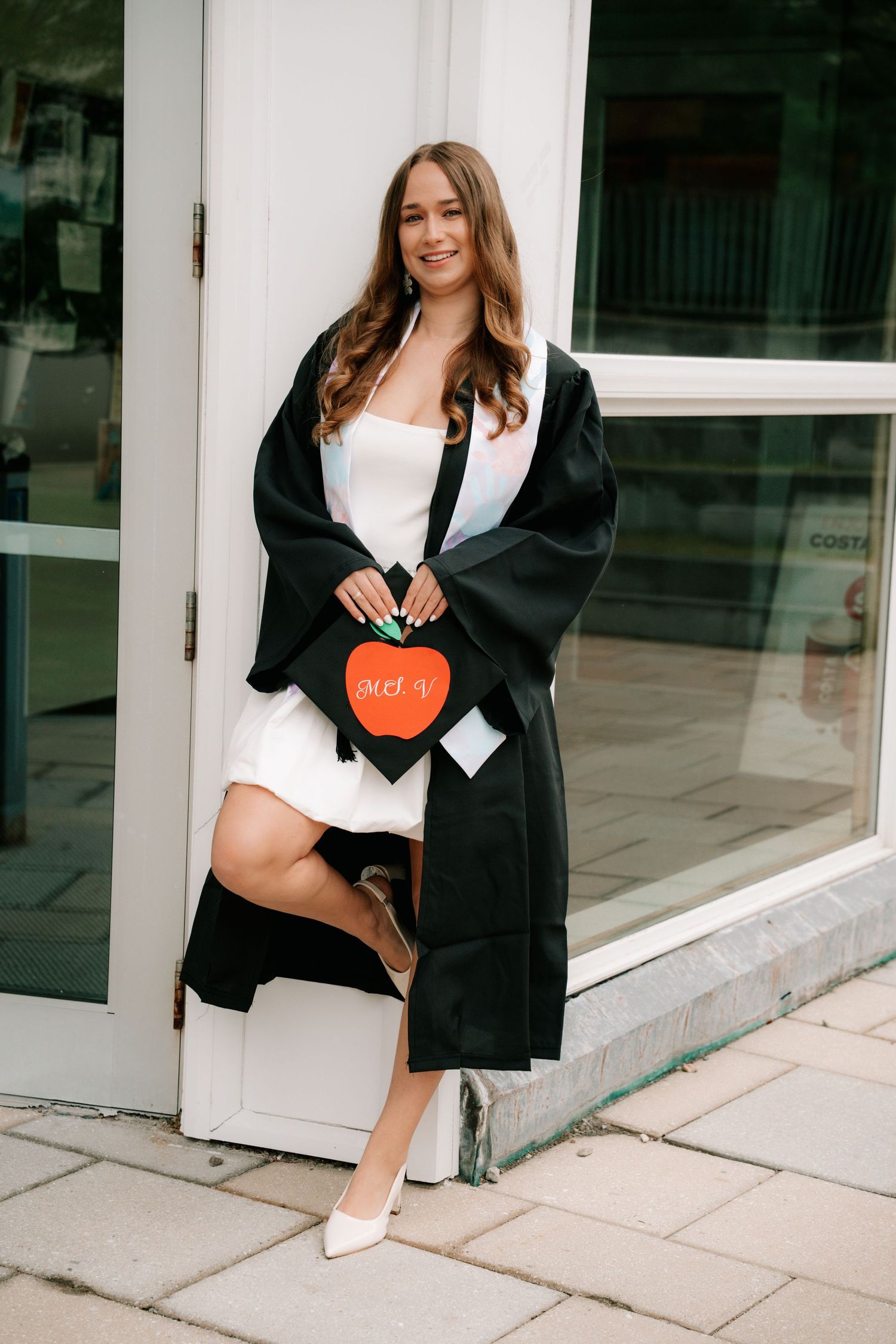 Female graduate in white dress holding decorated graduation cap with apple design outside building.
