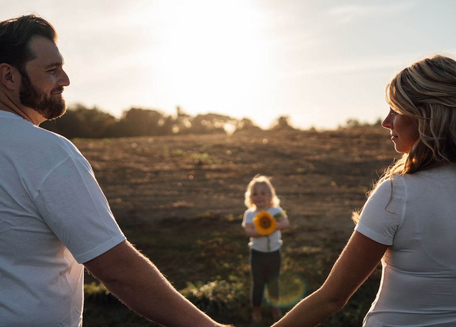 A couple holds hands smiling at each other while their young child stands in a sunlit open field holding a sunflower.